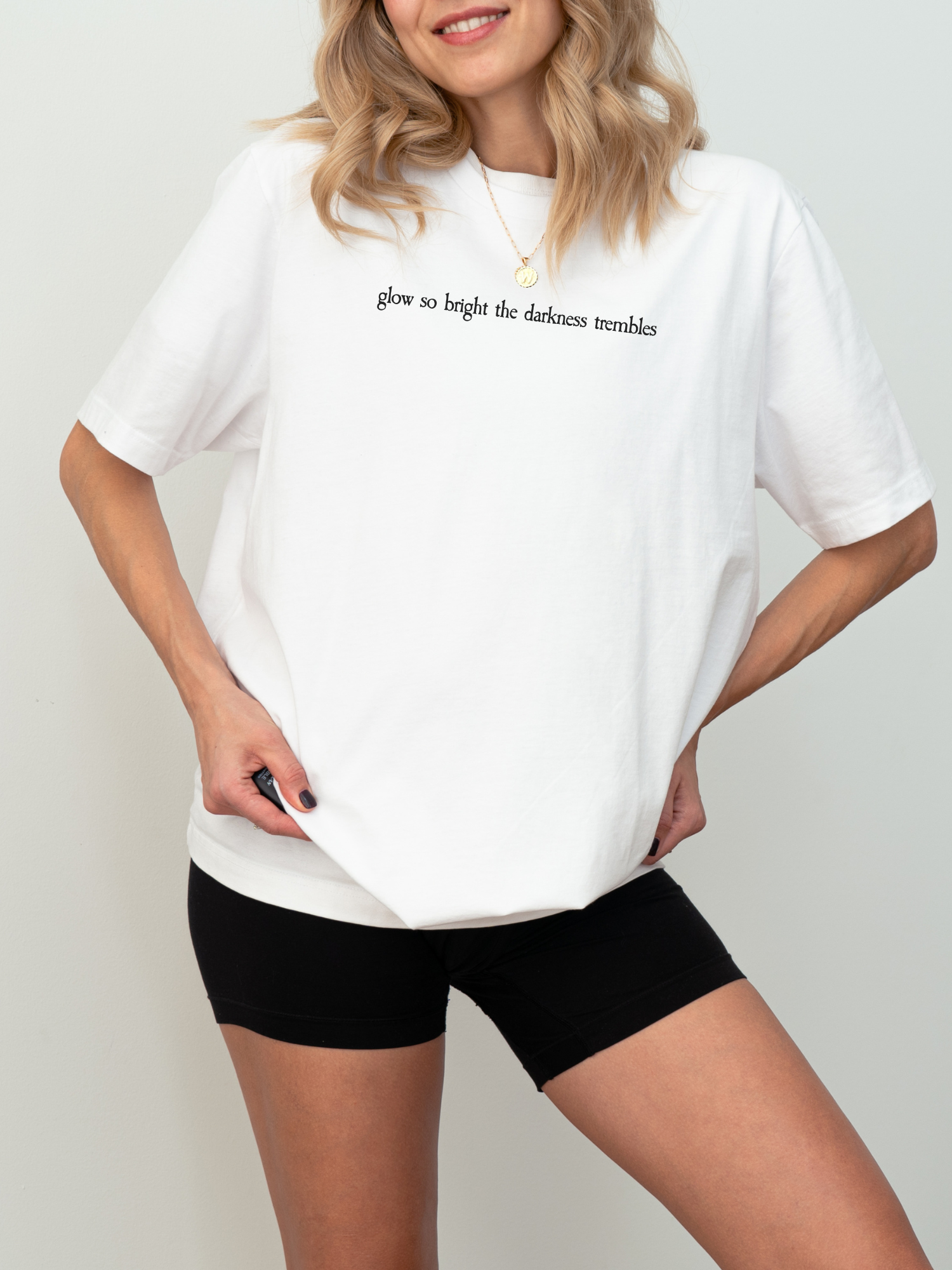 Bright white Bella Canvas tee featuring the fantasy quote “glow so bright the darkness trembles” in serif black font, styled simply with biker shorts and layered jewelry