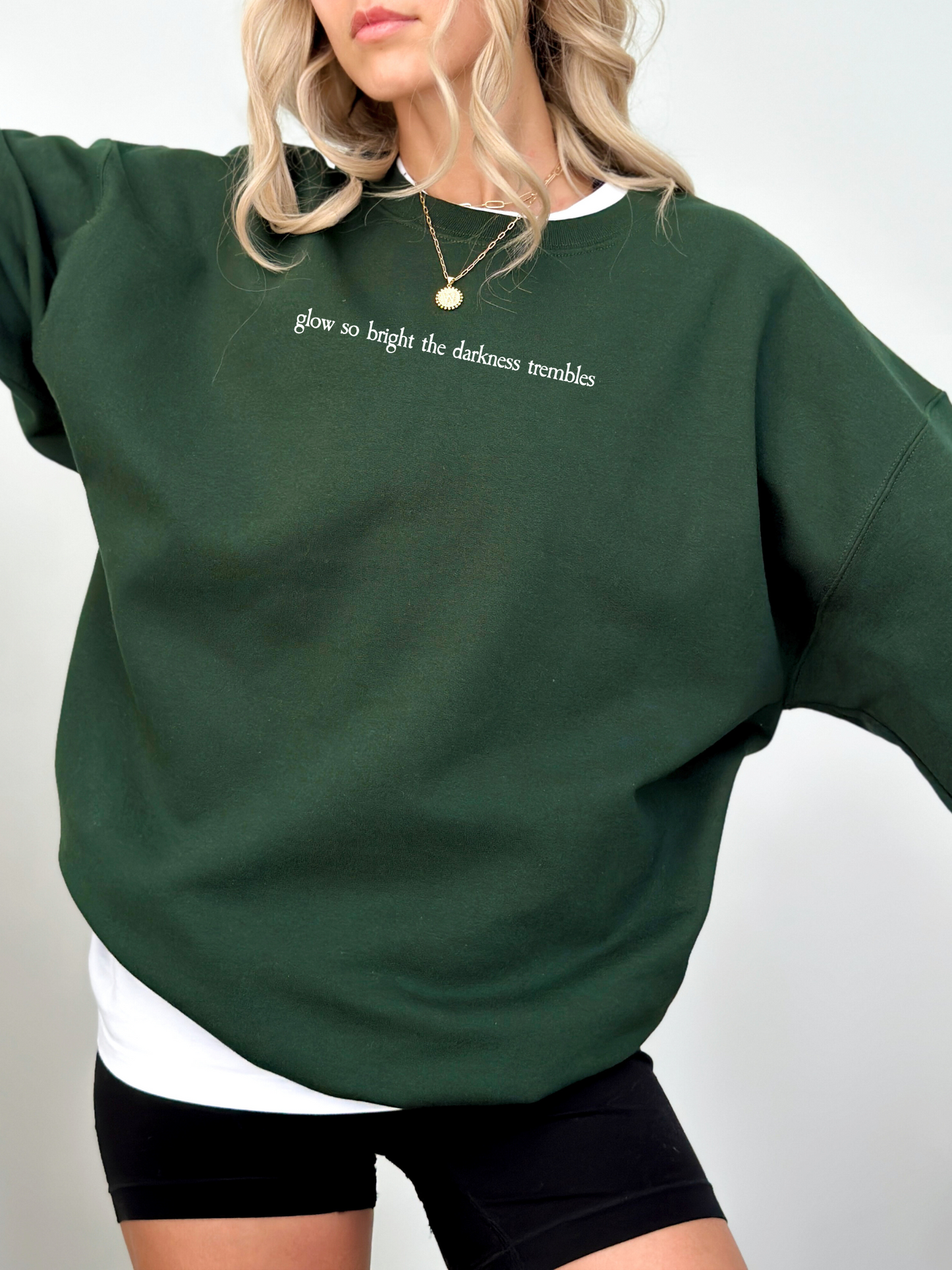 Model wearing a deep forest green Gildan sweatshirt with white serif quote across the chest. The text reads “glow so bright the darkness trembles.” She is styled with black biker shorts and a layered gold necklace.