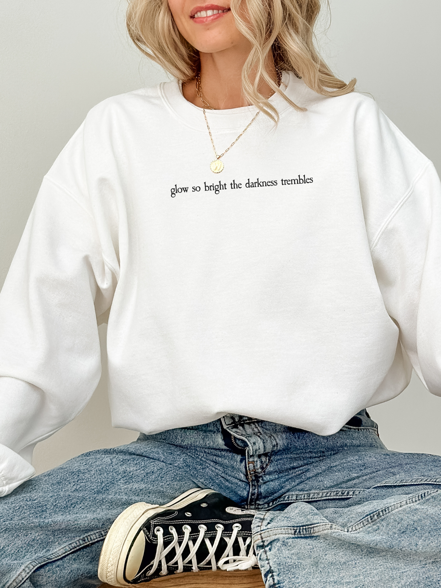 Bright white Gildan sweatshirt with elegant black serif quote "glow so bright the darkness trembles." Worn by a woman sitting cross-legged in jeans and black high-top sneakers, giving off a radiant, minimal look.