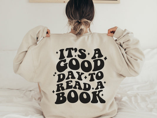 woman sitting on bed wearing sand-colored sweatshirt with large back print that says it's a good day to read a book