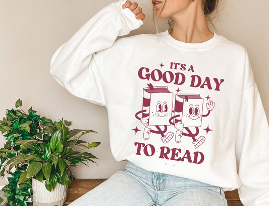 woman wearing white sweatshirt with it's a good day to read graphic, seated beside green plant