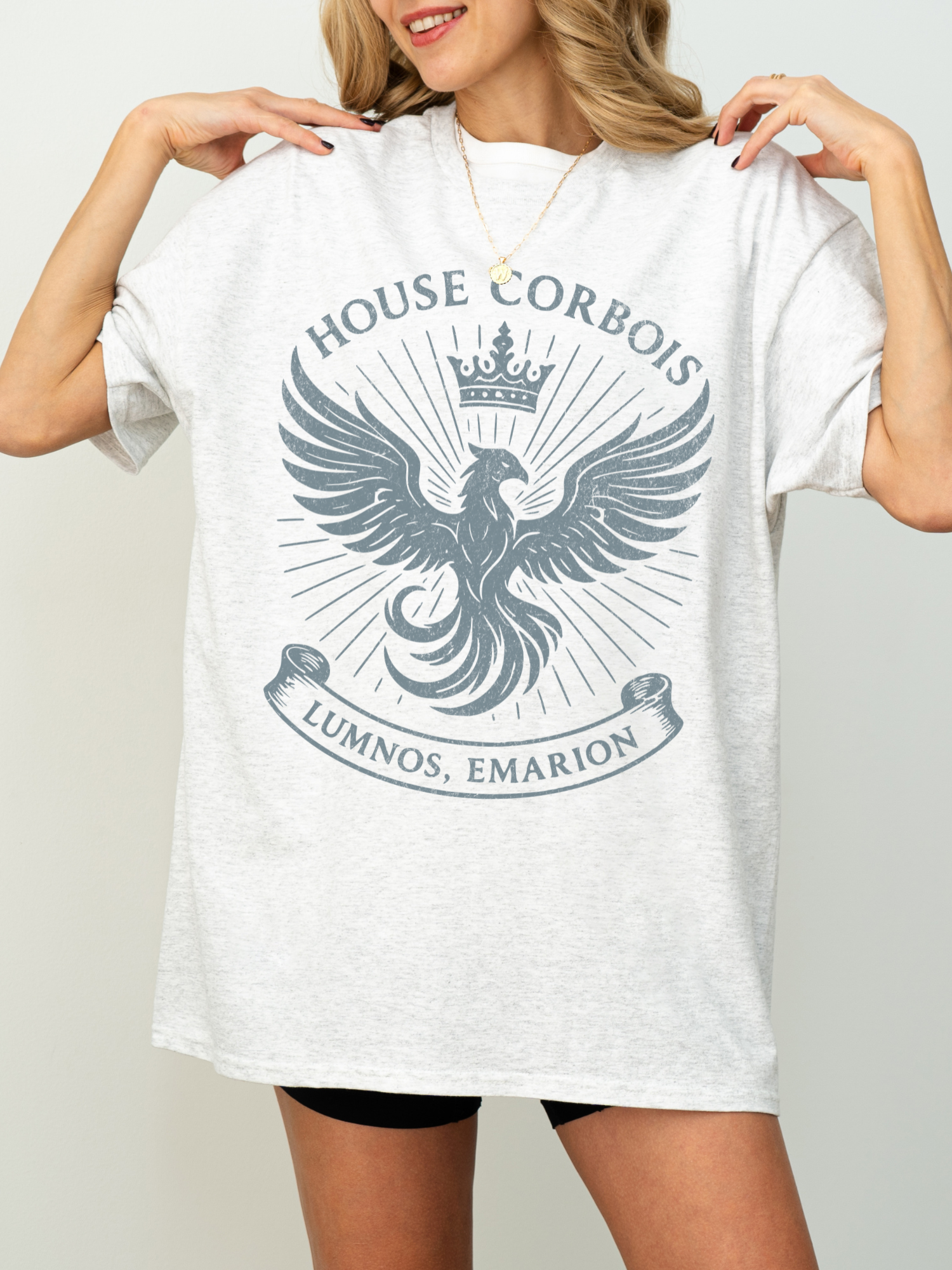 Model wearing a white House Corbois t-shirt featuring a large vintage-style phoenix crest with crown and rays, inspired by the realm of Luminos, Emarion. The shirt is styled casually with biker shorts for a comfy fantasy look.