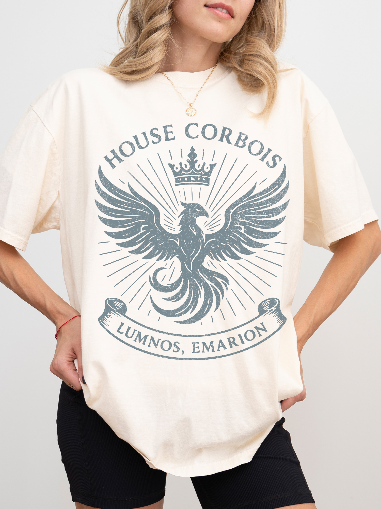 Model showcasing a natural-colored t-shirt with a detailed blue-grey phoenix crest, banner, and crown design from the House Corbois of Luminos, Emarion. Styled for casual, cozy vibes for book nerds and fantasy readers.