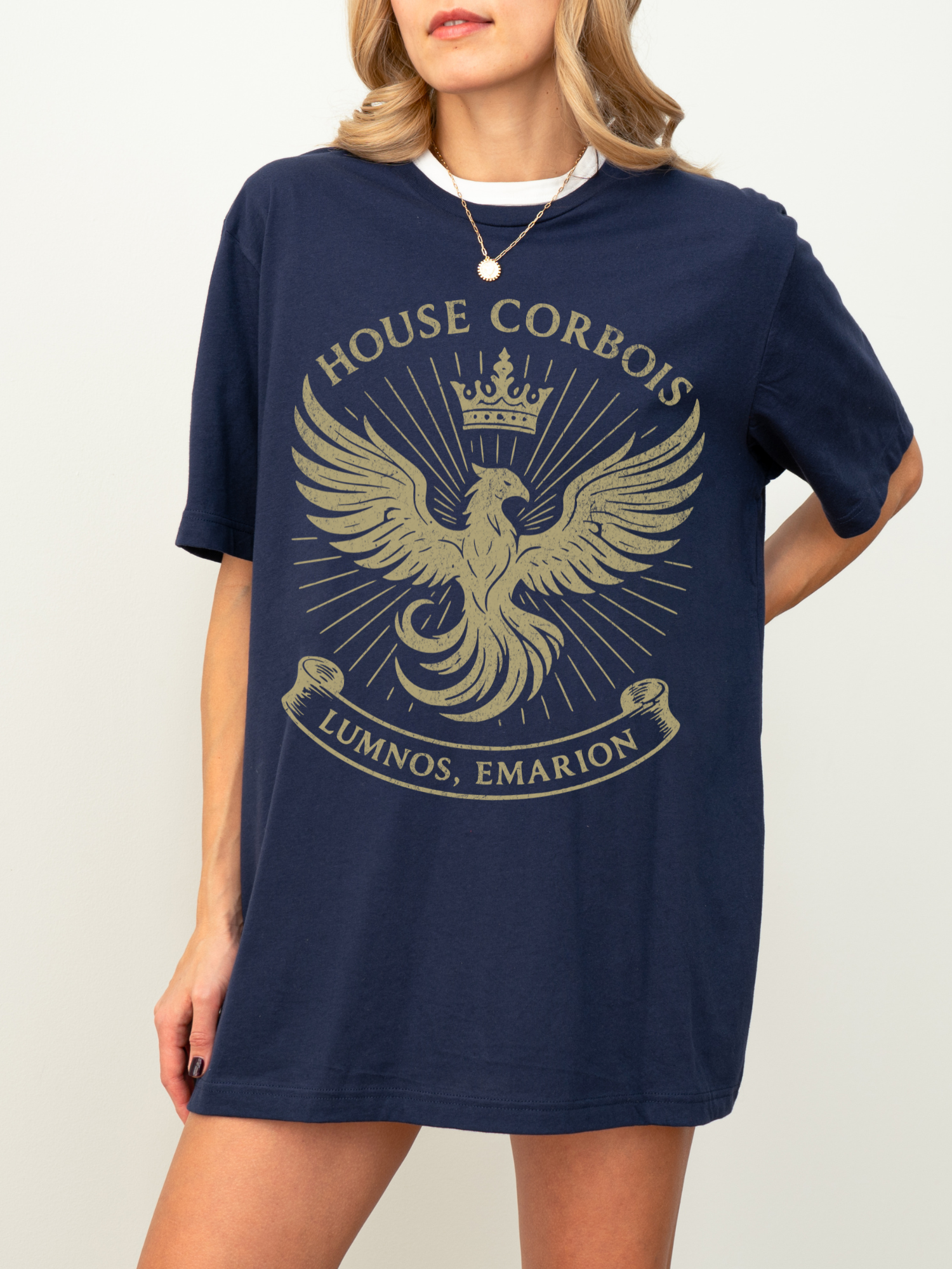Model in navy shirt featuring a large golden House Corbois phoenix emblem with crown and swirling rays. Styled as an oversized tee dress for a powerful and stylish fantasy reader look.