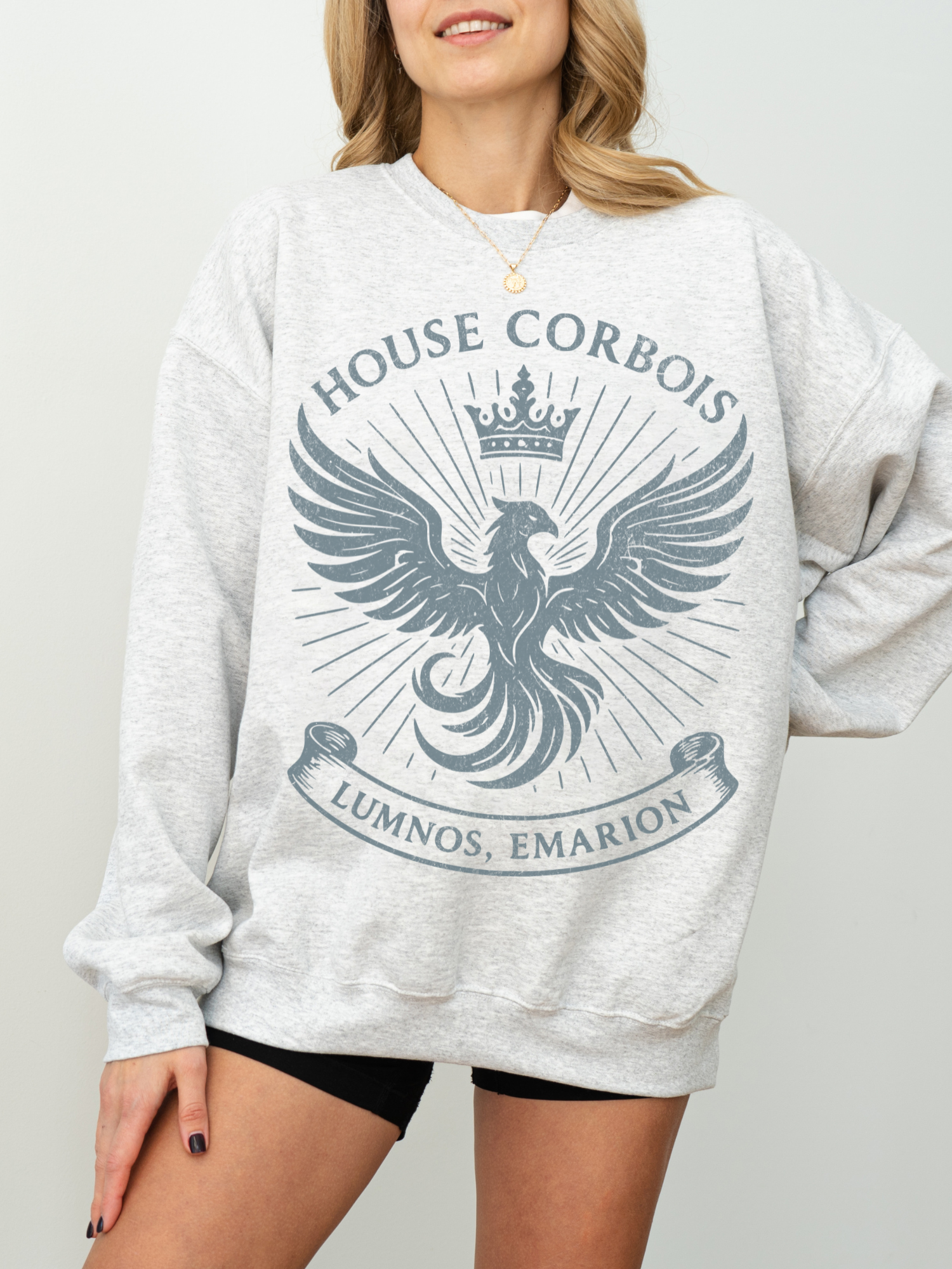Woman wearing light grey crewneck with blue bookish phoenix design and crown above text