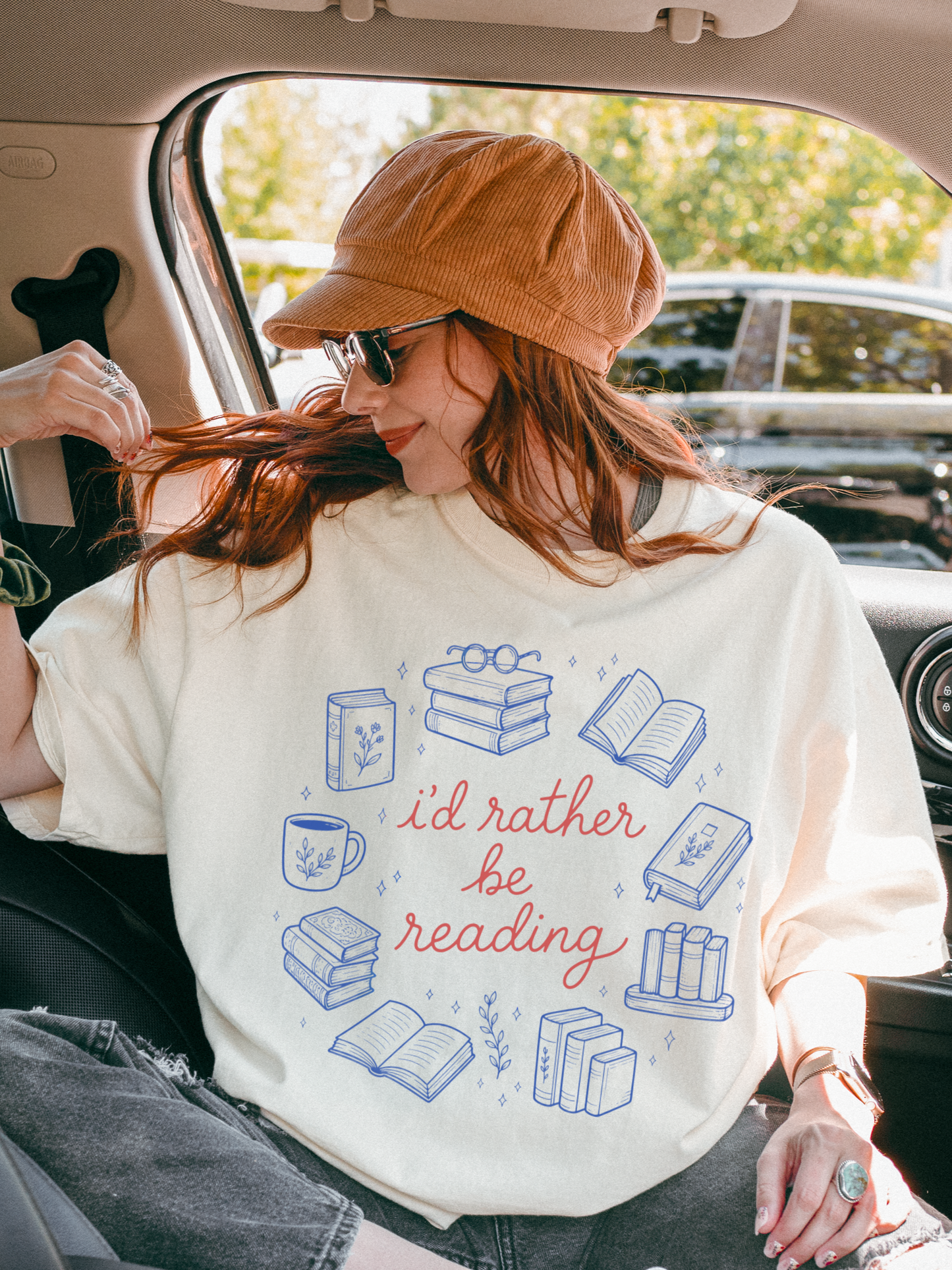 woman sitting in car wearing oversized natural cream bookish graphic shirt with id rather be reading design and illustrated books coffee mug and reading icons