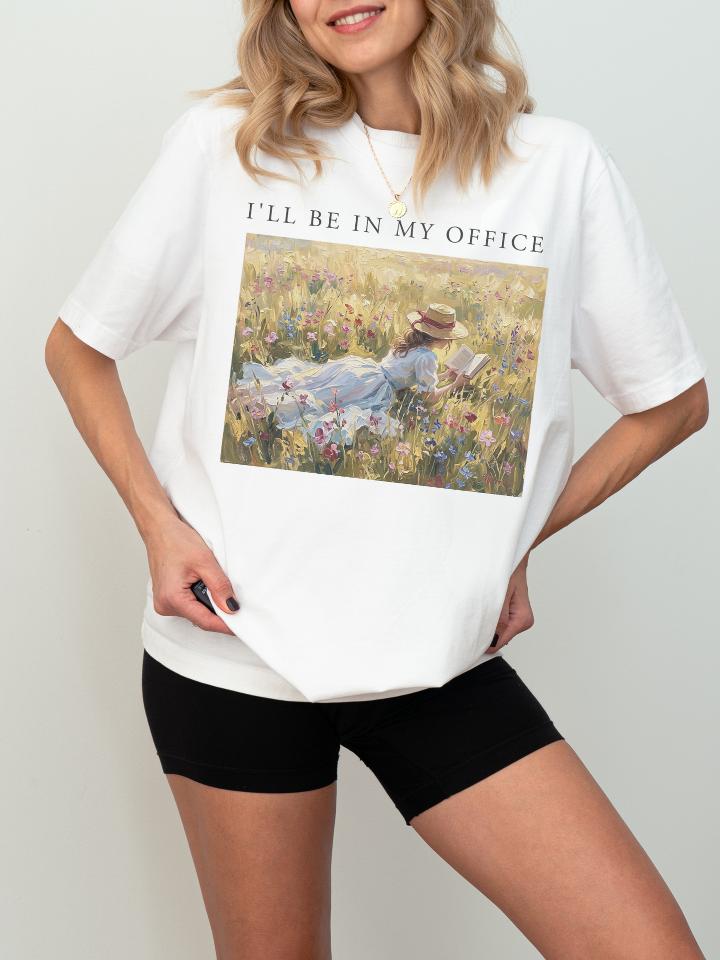 Woman wearing a white t shirt with I’ll be in my office text and a romantic impressionist artwork of a girl reading in a sunlit wildflower field