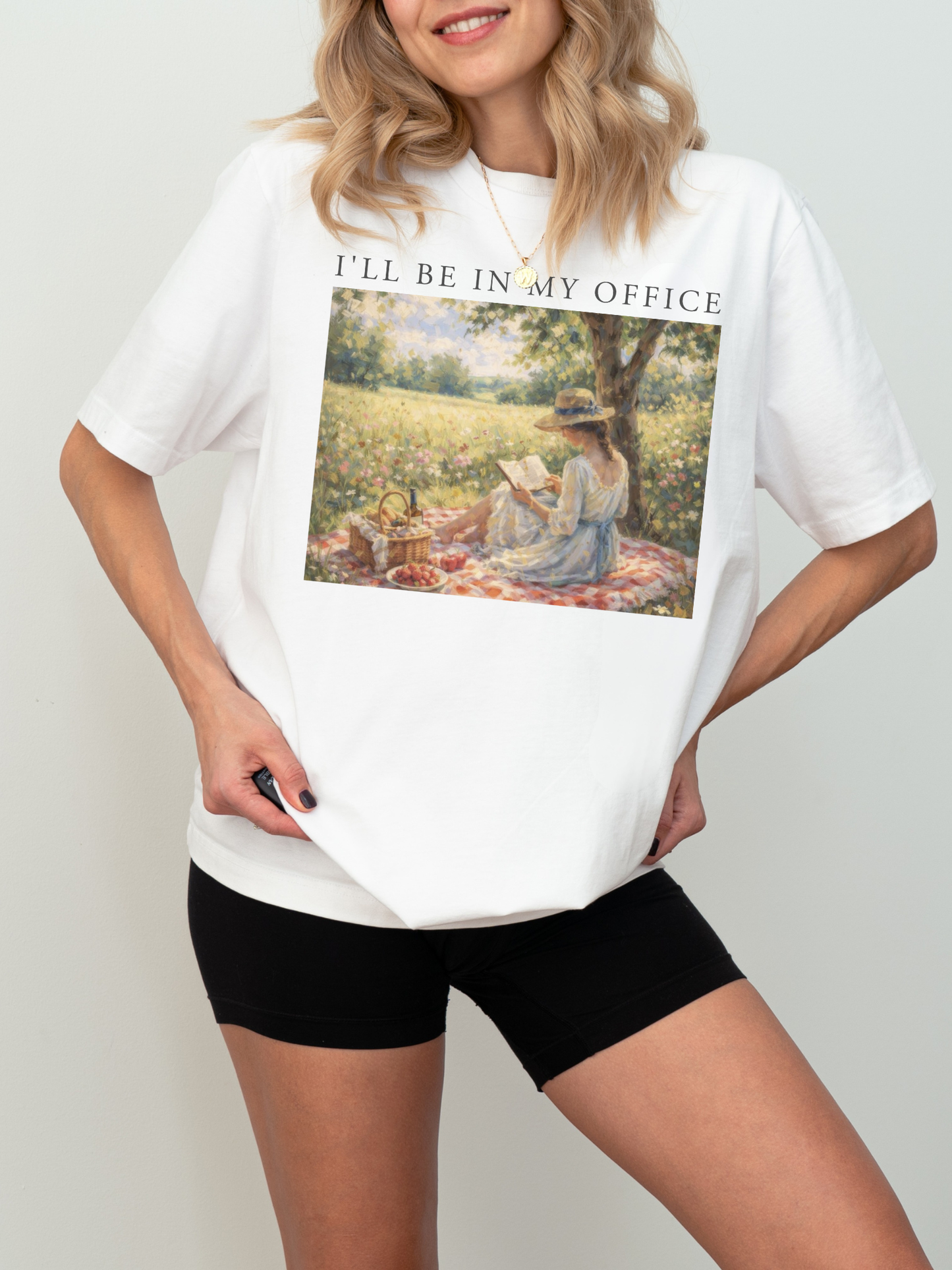 Woman wearing a white t shirt with I’ll be in my office text and a romantic vintage painting of a woman reading a book in a sunlit meadow picnic setting