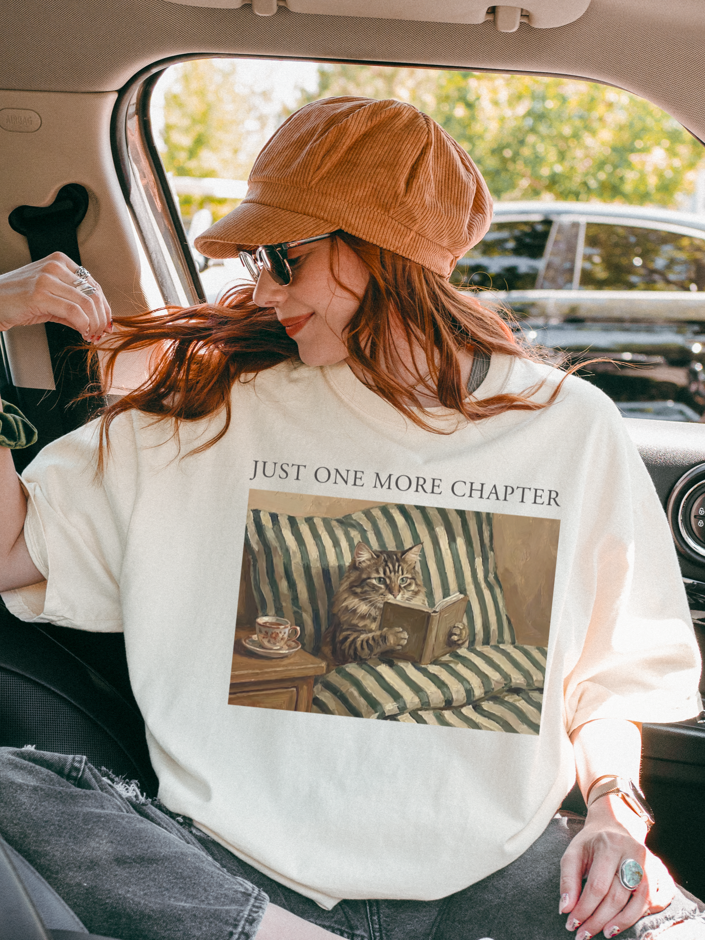 Woman sitting in a car wearing an ivory t shirt with just one more chapter text and a cozy cat reading a book illustration on a striped couch
