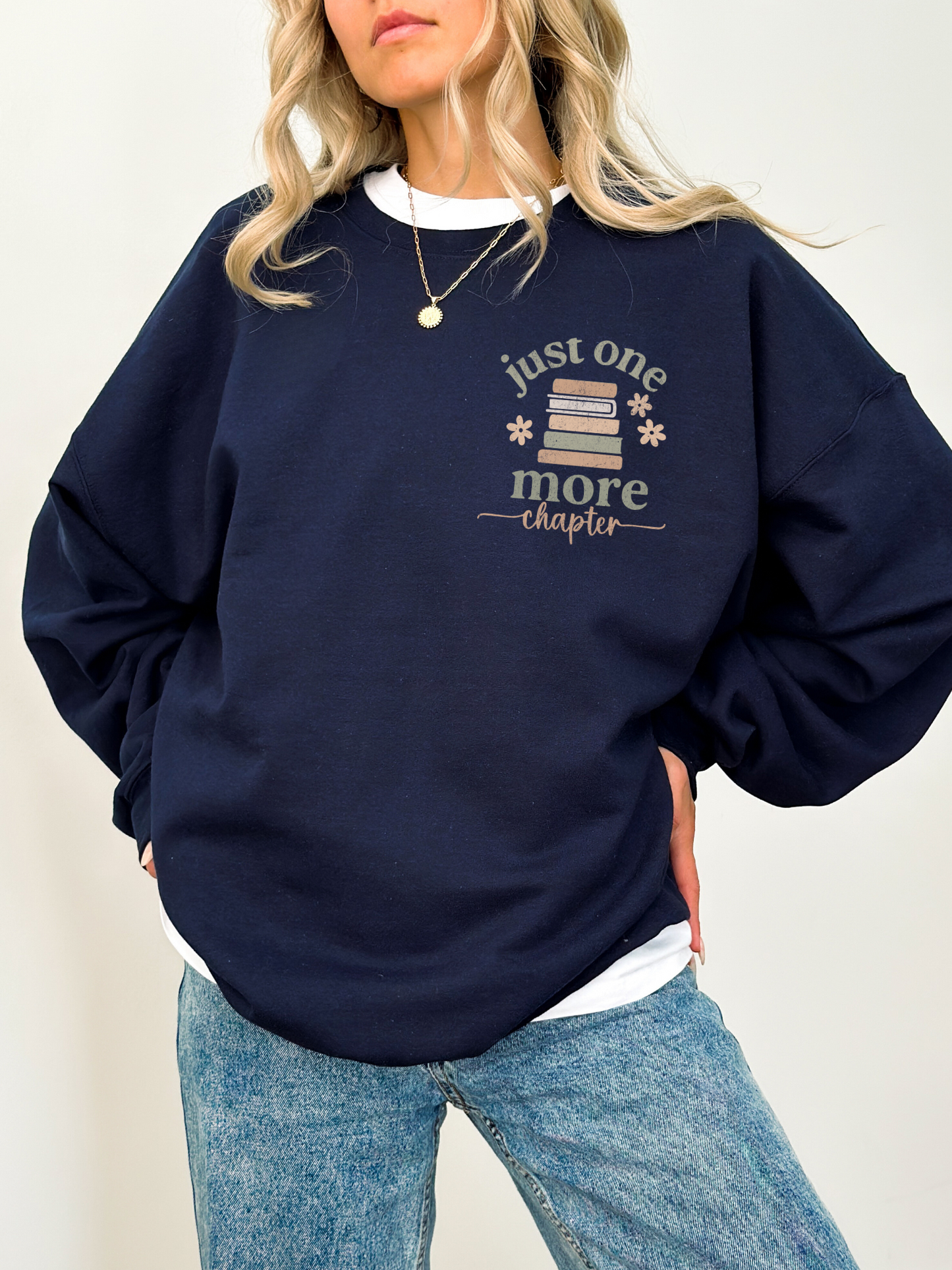 Woman in a navy crewneck sweatshirt featuring a front left chest design with stacked vintage-style books, soft florals, and just one more chapter in warm neutral lettering. Styled with light blue jeans and white tee underneath.