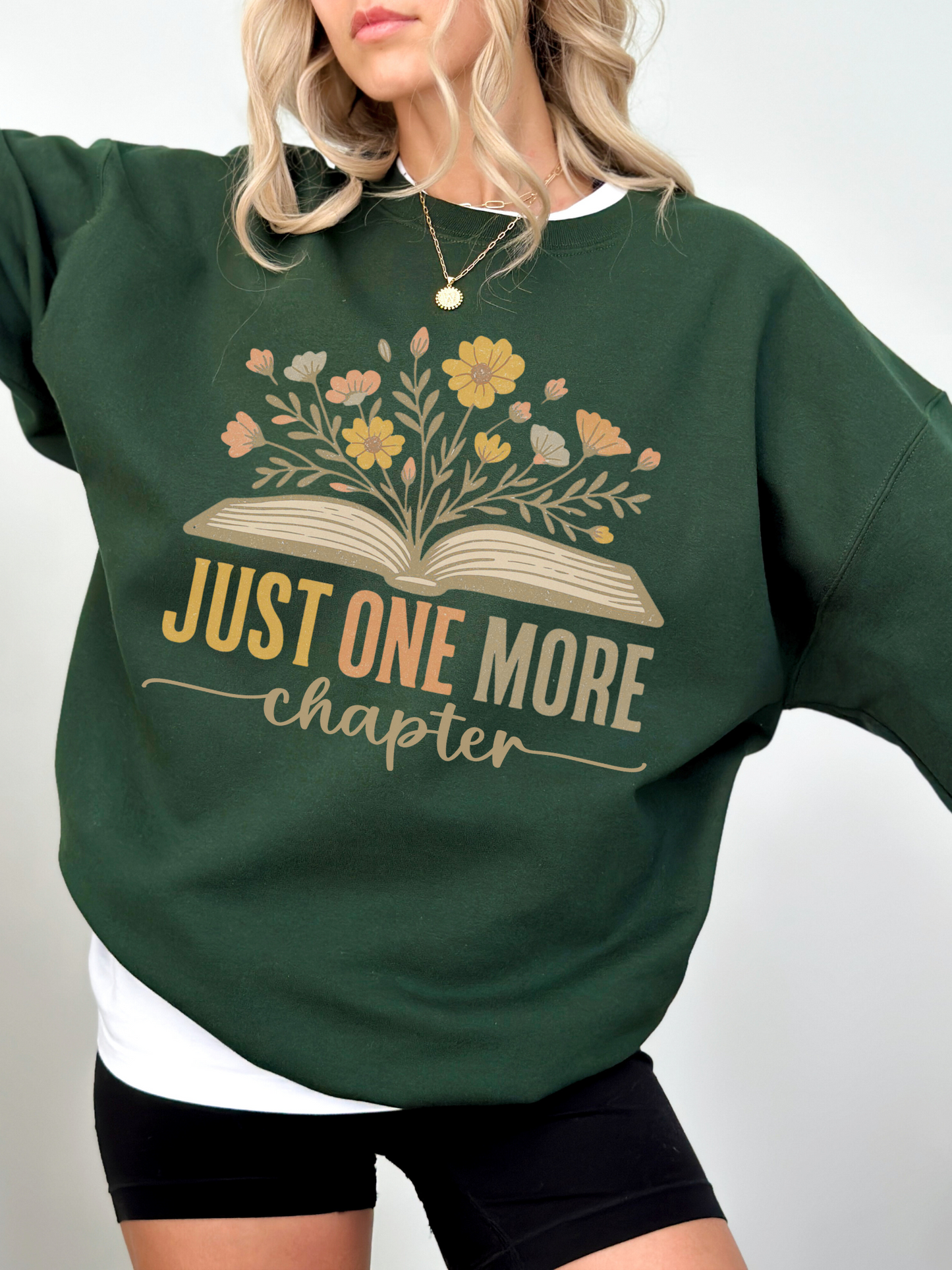 Woman in forest green cozy sweatshirt featuring vintage-style floral book graphic and just one more chapter quote, layered over a white shirt and styled with black shorts, standing with hand on hip