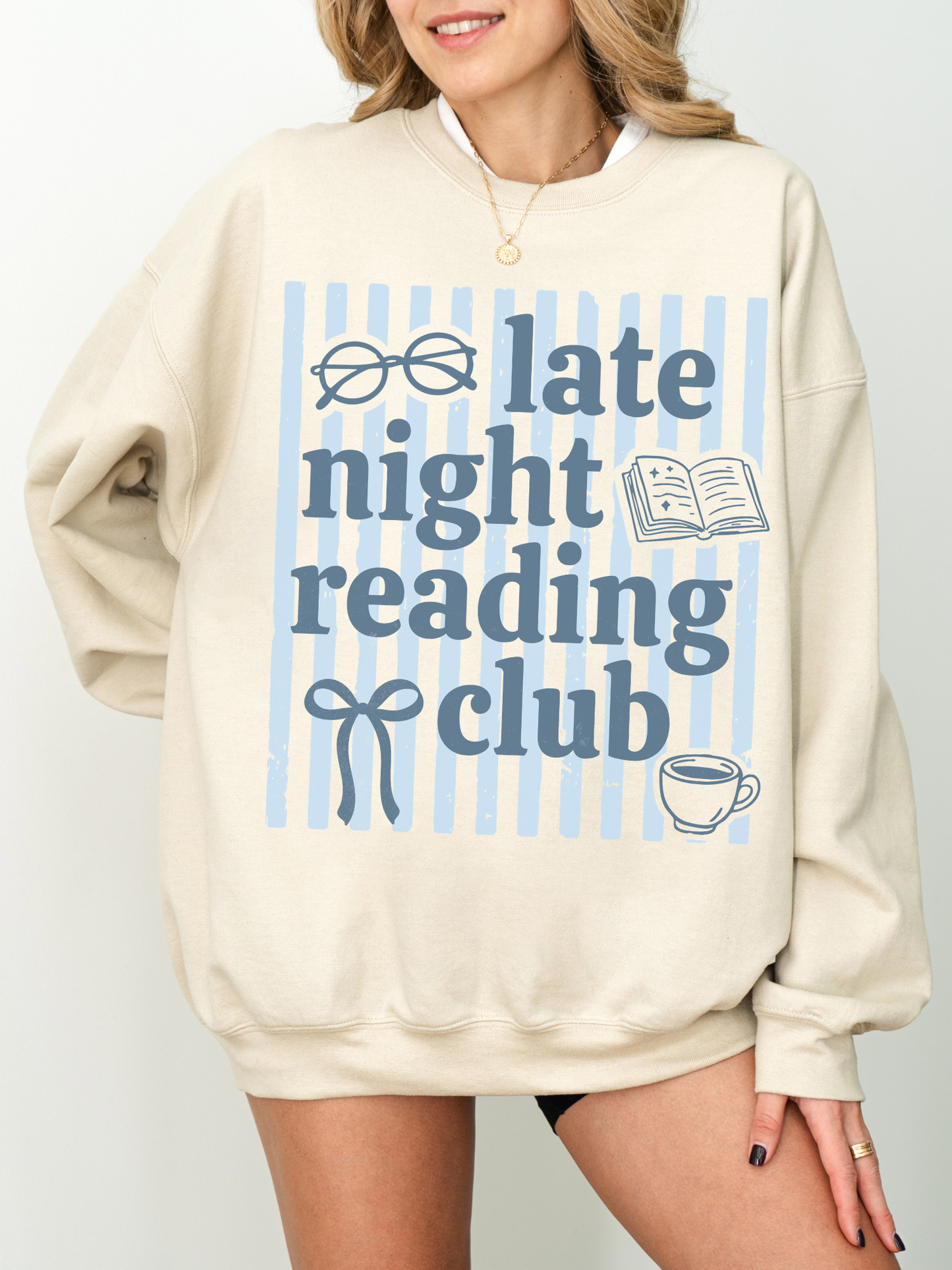 lifestyle photo of woman in sand sweatshirt with late night reading club design featuring glasses, open book, coffee cup and soft blue stripes