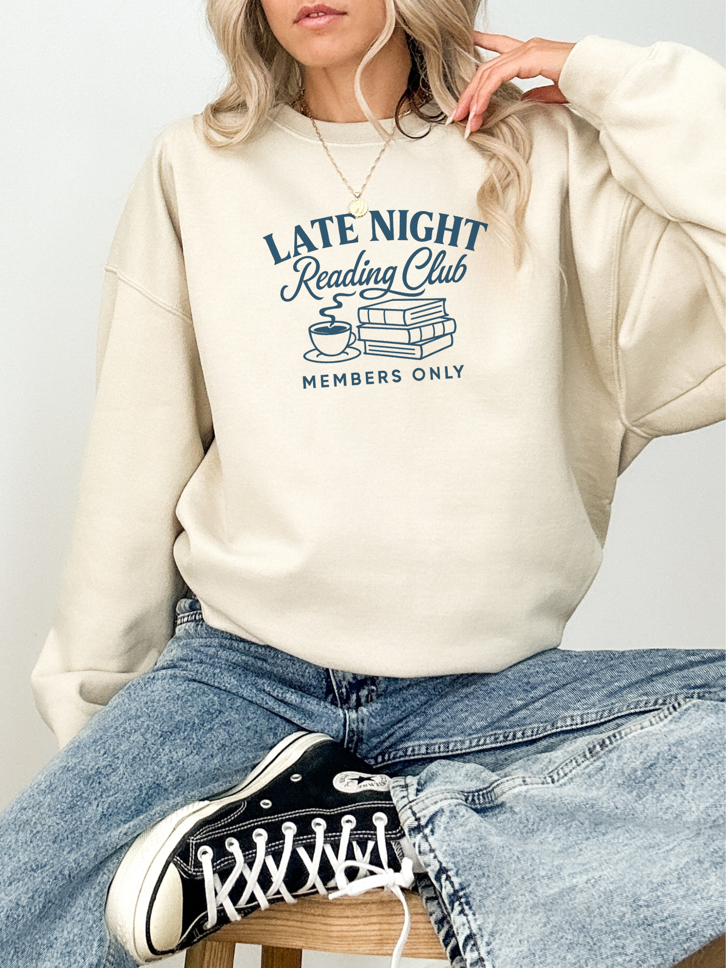 woman sitting cross-legged in a sand sweatshirt with late night reading club members only and navy blue graphic