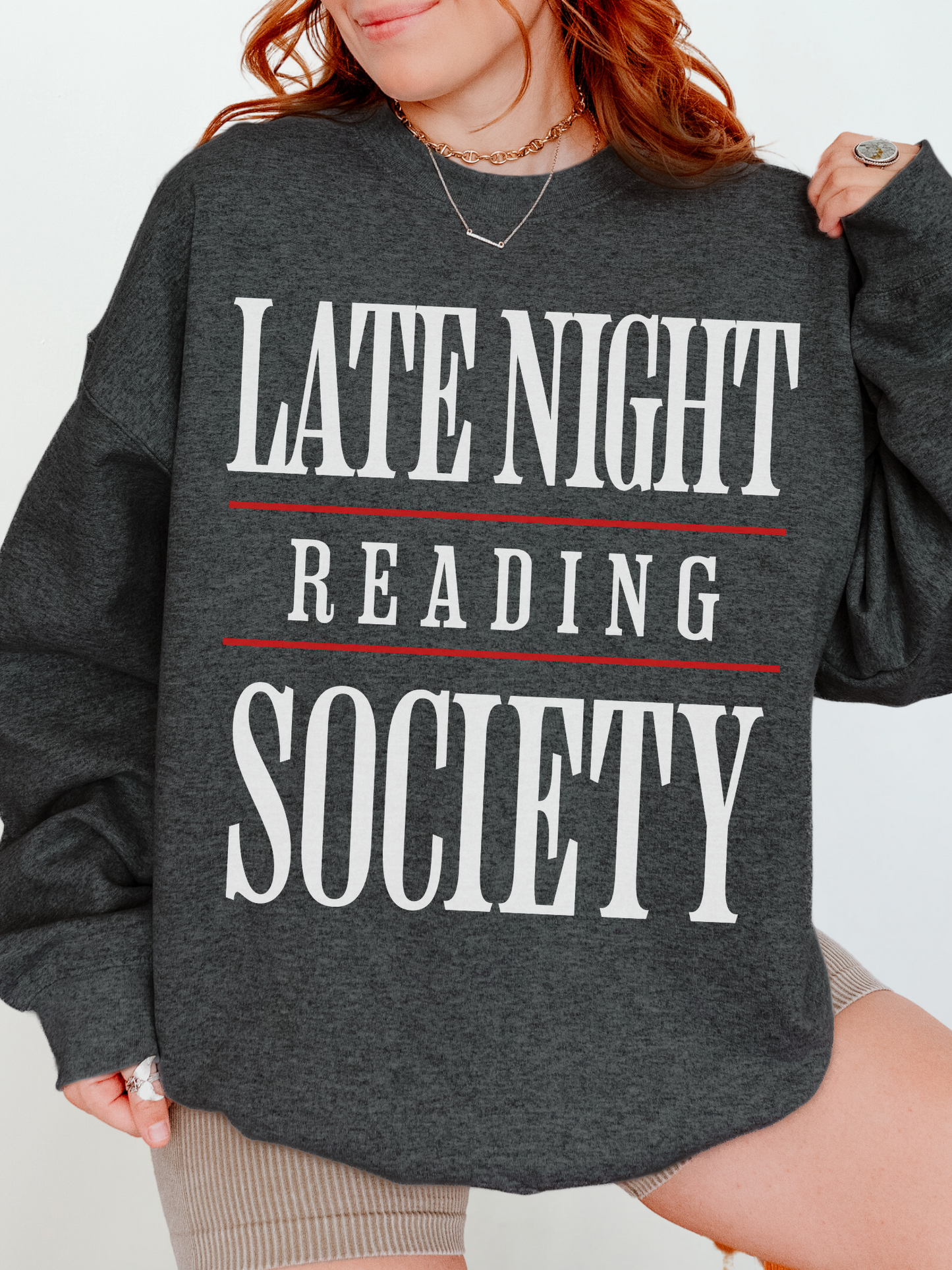 woman wearing a dark heather grey sweatshirt with large white and red graphic text that reads late night reading society