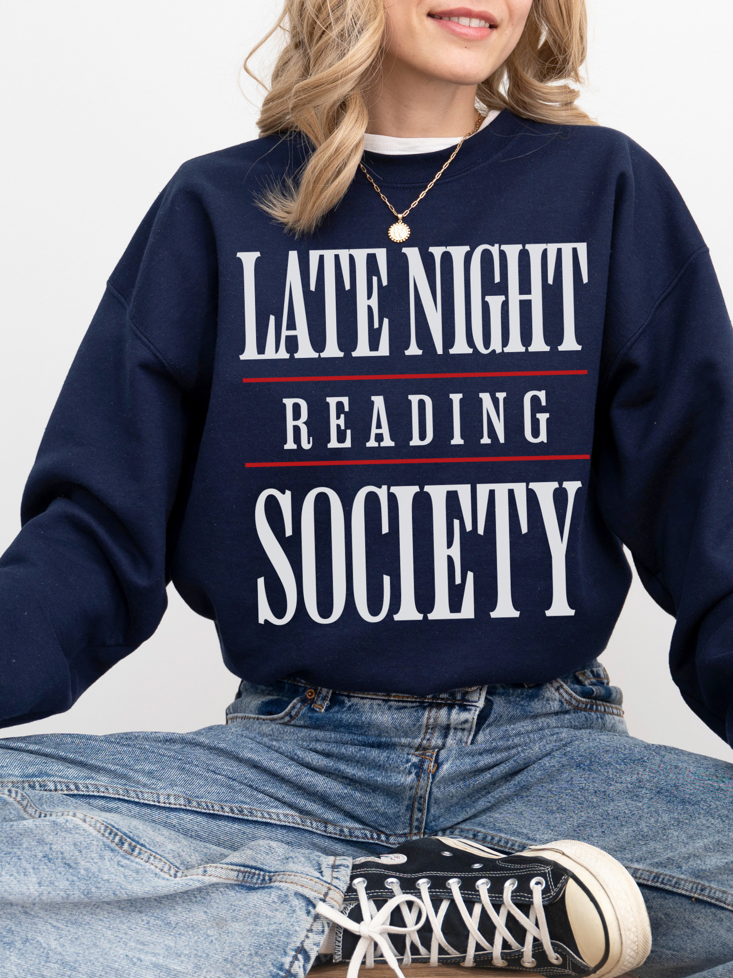 woman sitting in a navy sweatshirt with bold white and red text that says late night reading society