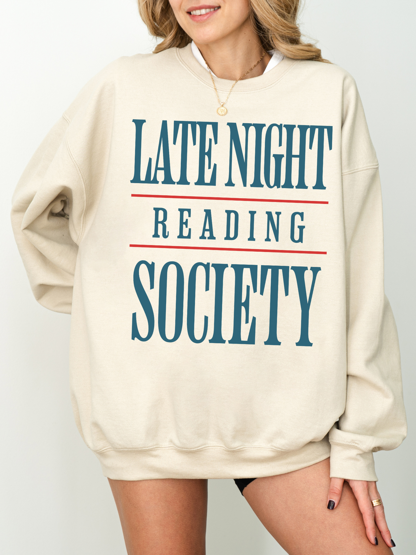 woman wearing a sand crewneck sweatshirt with large blue and red text that says late night reading society