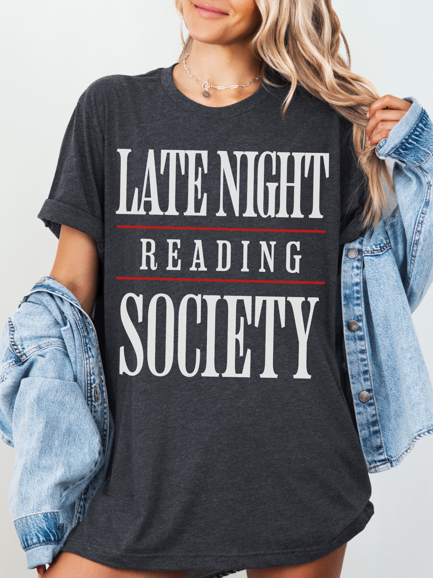 woman wearing a dark heather grey tee that says late night reading society in bold white font with red lines and denim jacket
