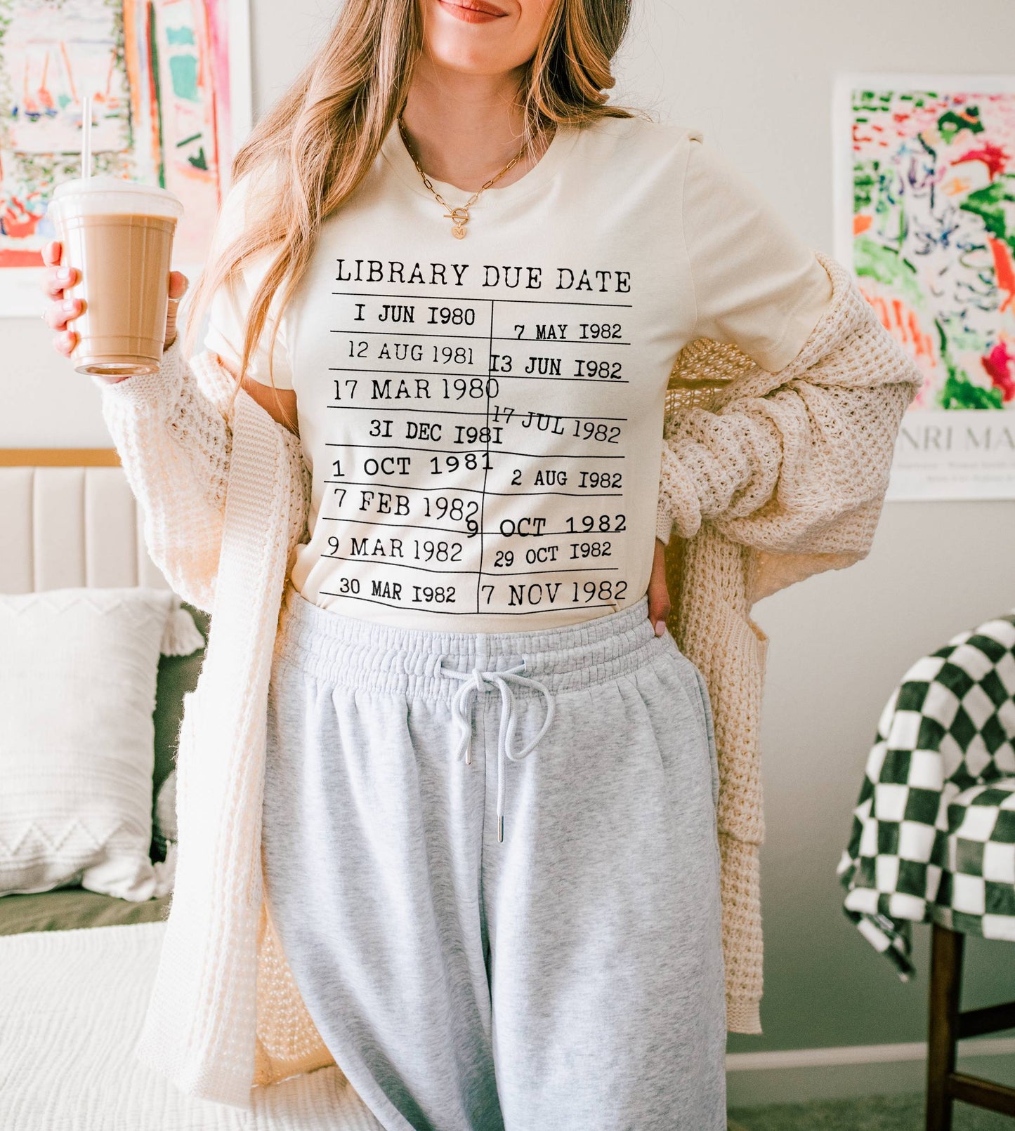 woman wearing a natural color t-shirt with vintage library due date stamp design, holding iced coffee