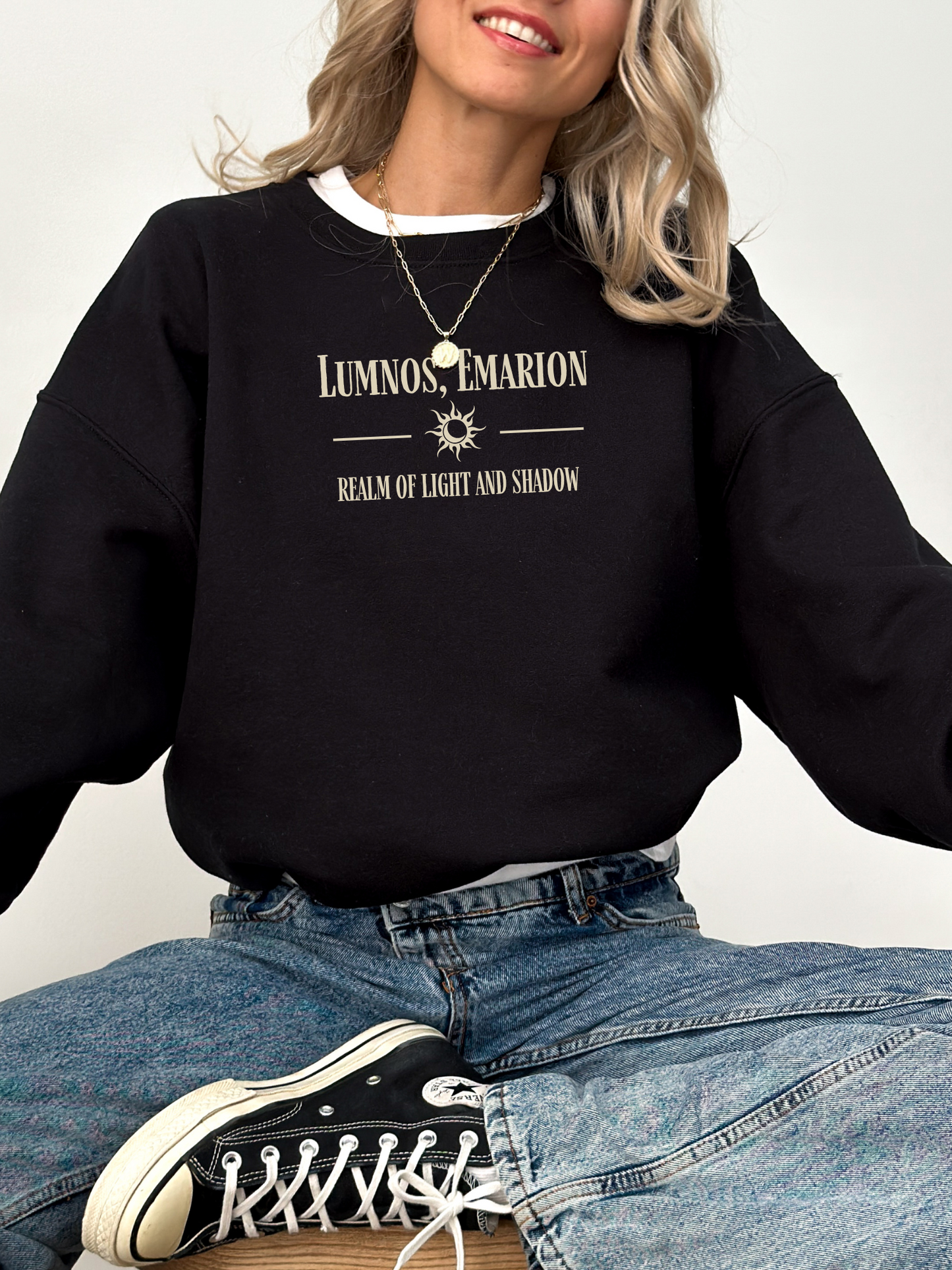 Woman sitting cross-legged in a black Gildan sweatshirt with “Lumnos, Emarion – Realm of Light and Shadow” quote, styled with jeans and layered necklaces.