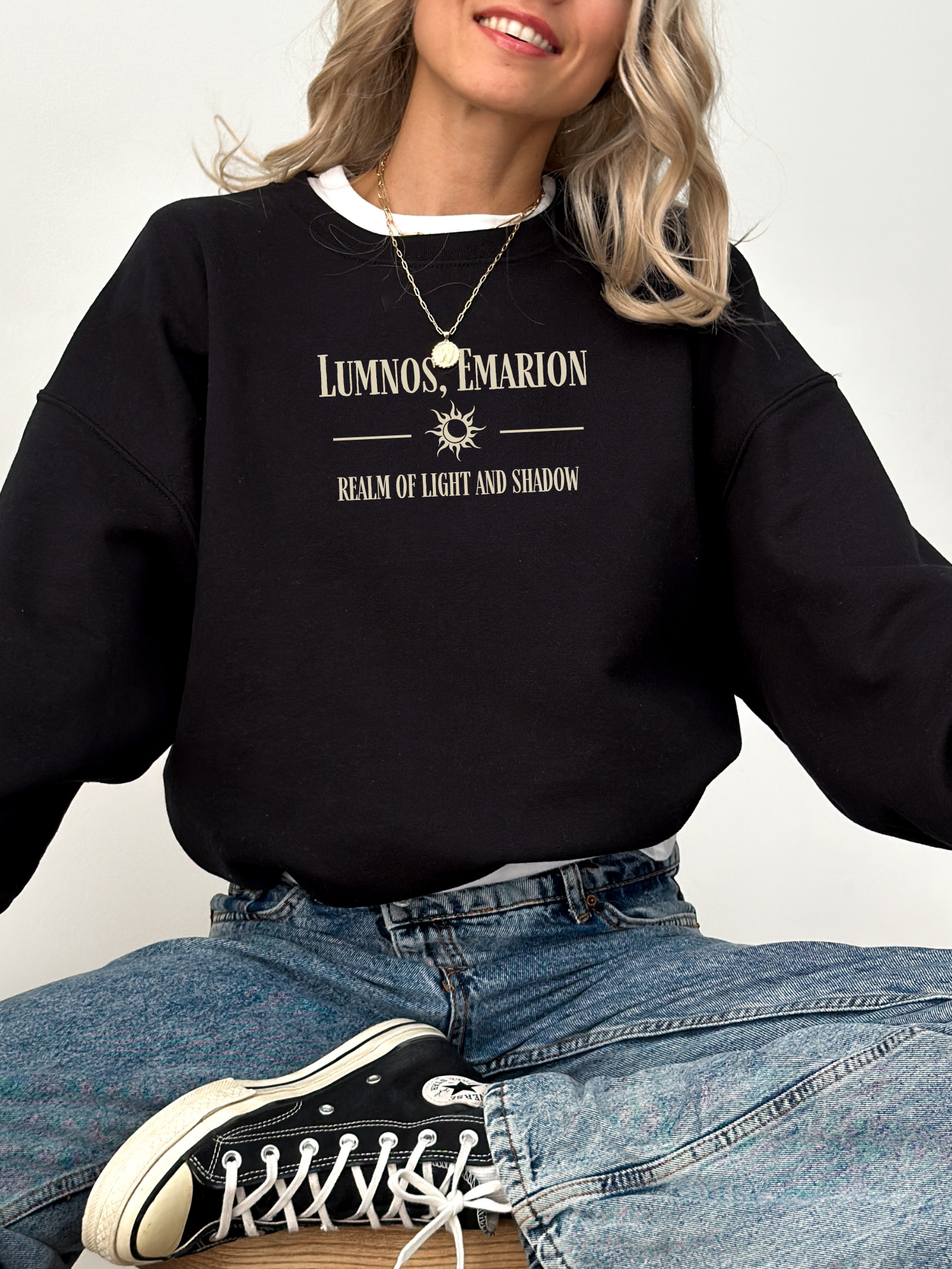 Woman sitting cross-legged in a black Gildan sweatshirt with “Lumnos, Emarion – Realm of Light and Shadow” quote, styled with jeans and layered necklaces.