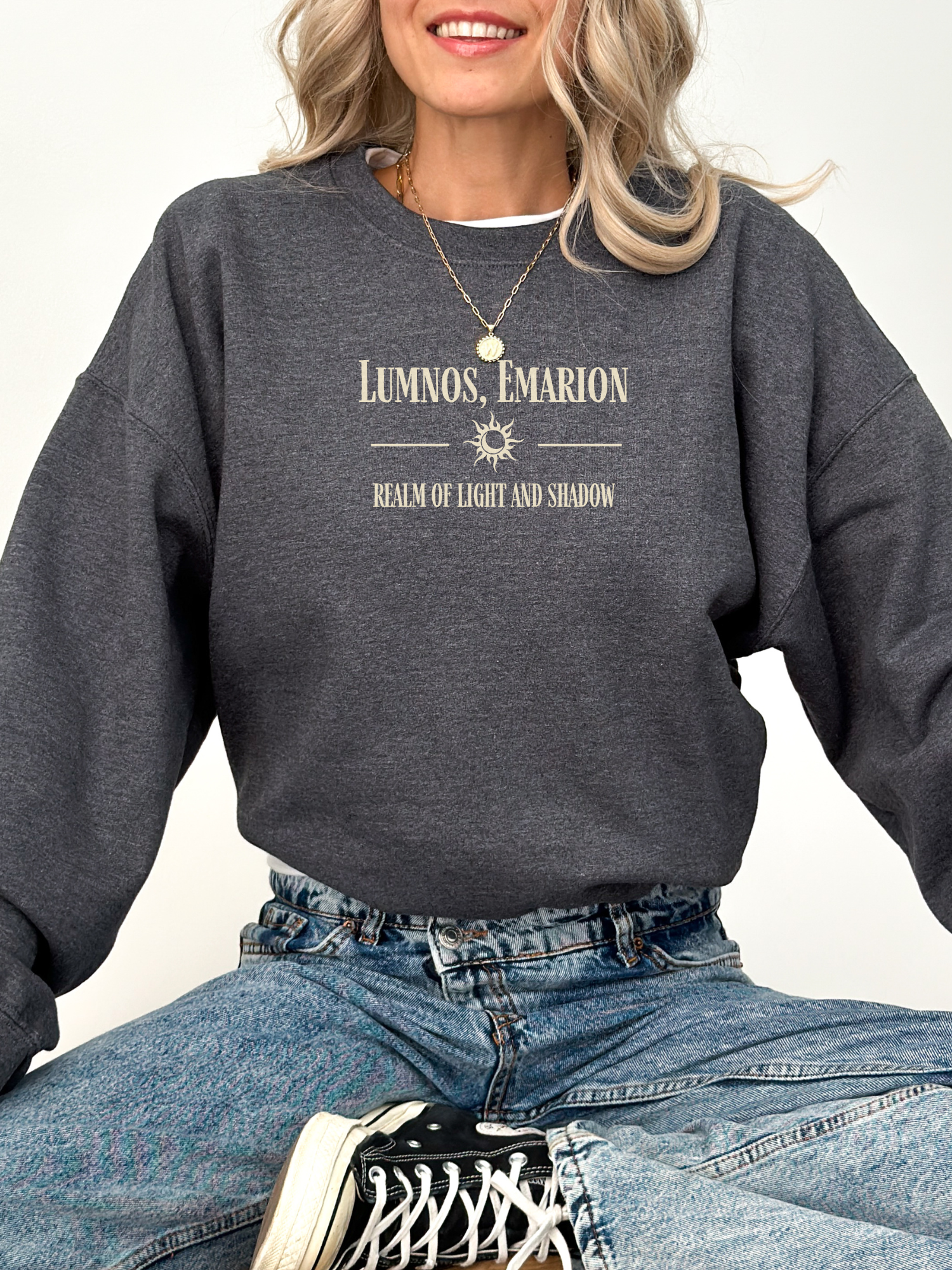 Woman smiling in a dark heather grey Gildan sweatshirt featuring “Lumnos, Emarion – Realm of Light and Shadow” design, paired with blue jeans and sneakers.
