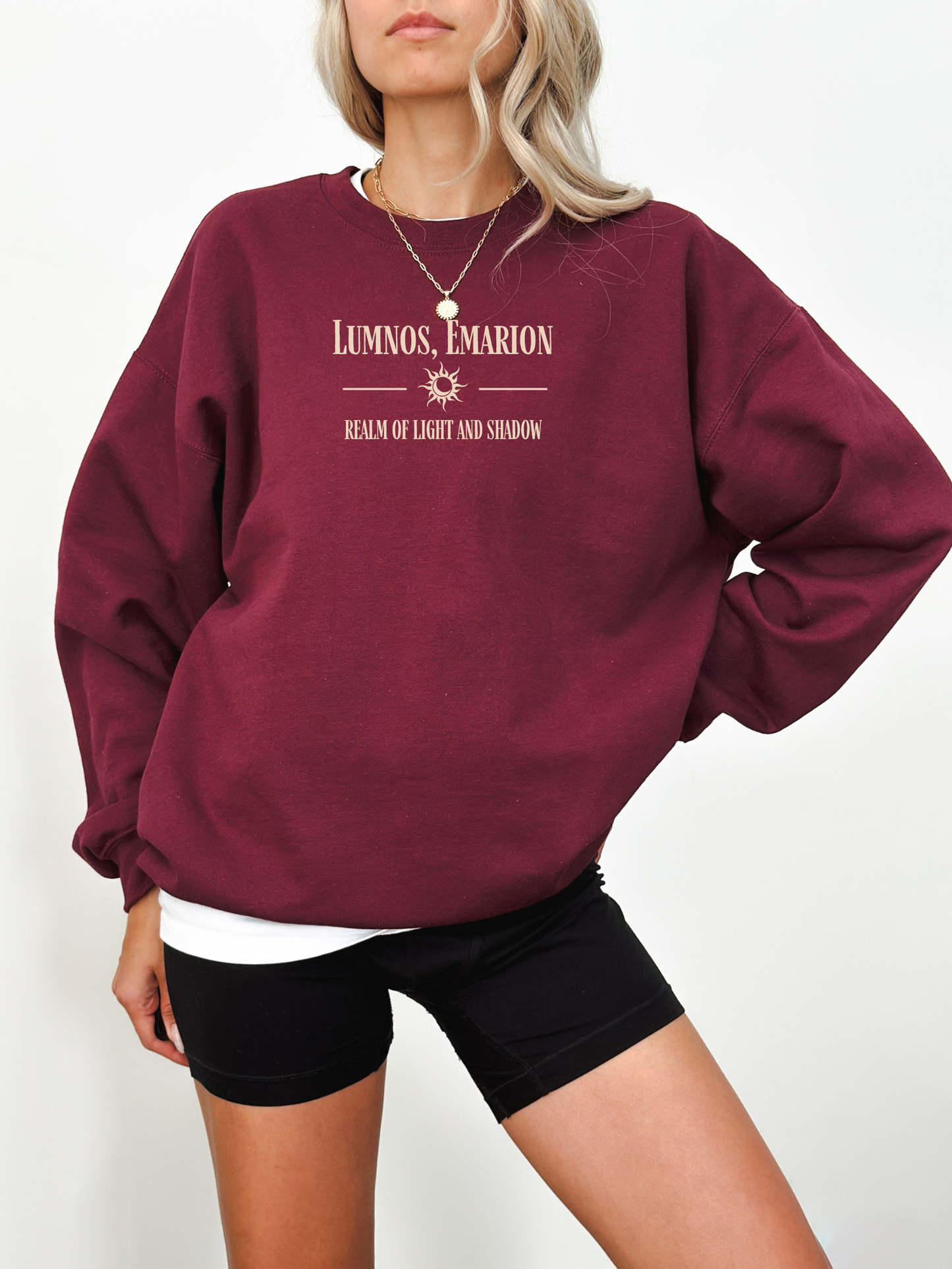 Woman wearing a maroon fantasy-themed Gildan sweatshirt reading “Lumnos, Emarion – Realm of Light and Shadow,” with a relaxed, cozy aesthetic.