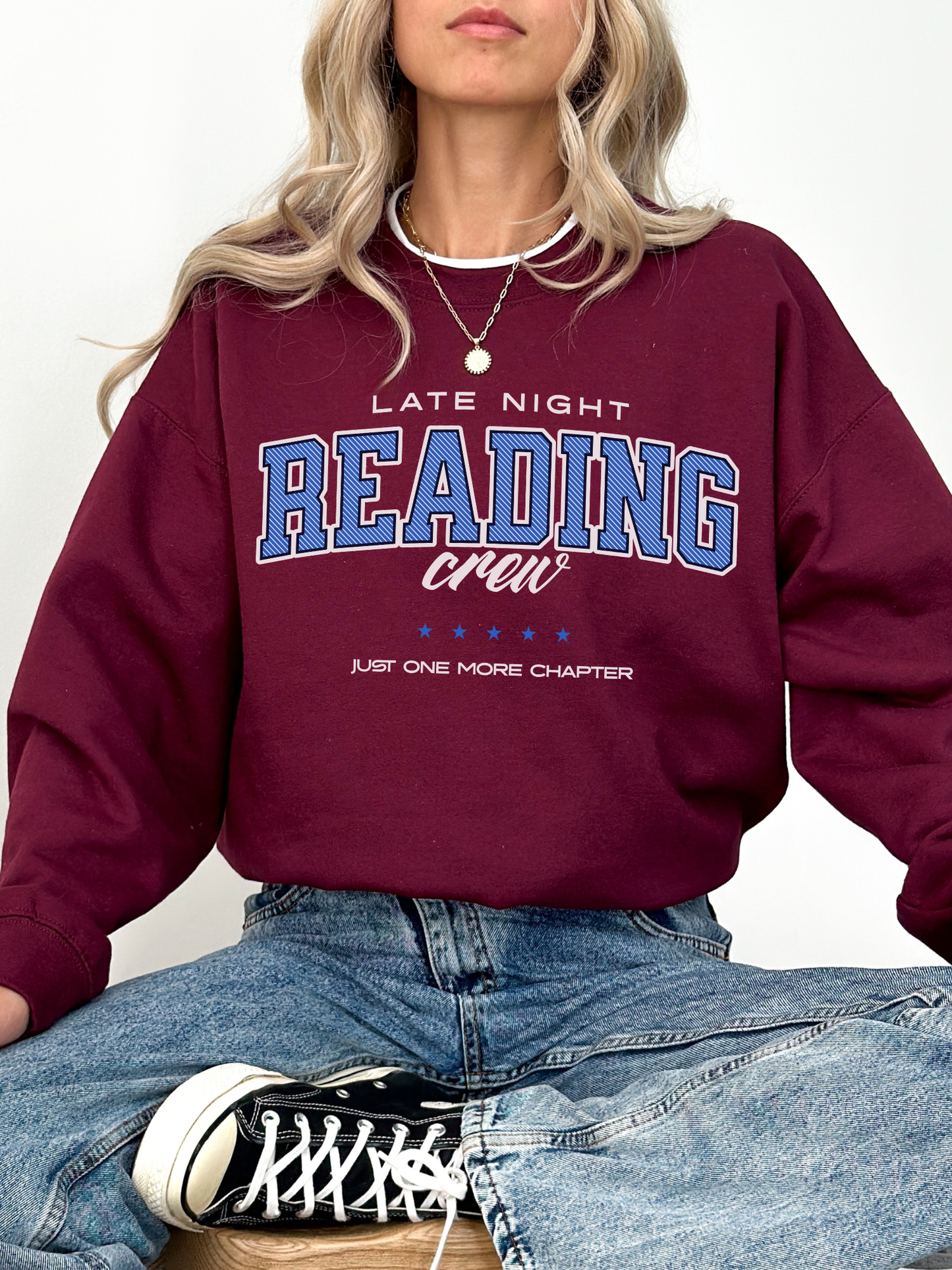 Maroon crewneck sweatshirt with bold “Late Night Reading Crew” text and stars, worn by woman sitting cross-legged in jeans