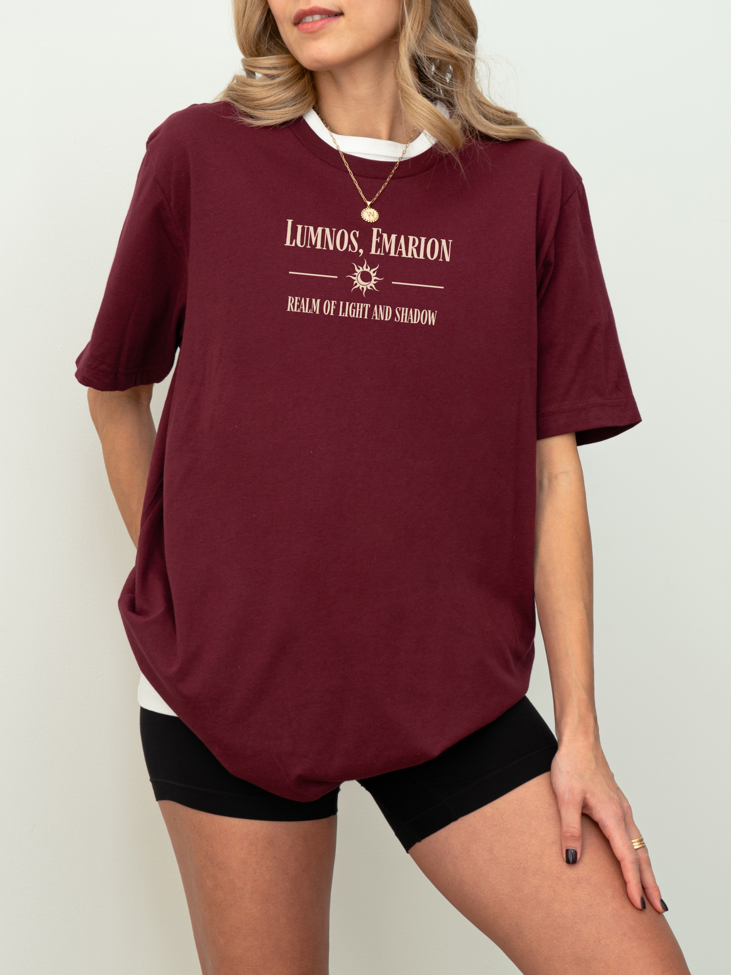 Model standing in maroon red shirt with cream fantasy text "Lumnos, Emarion - Realm of Light and Shadow", paired with black shorts and gold necklace, perfect for fantasy book lovers