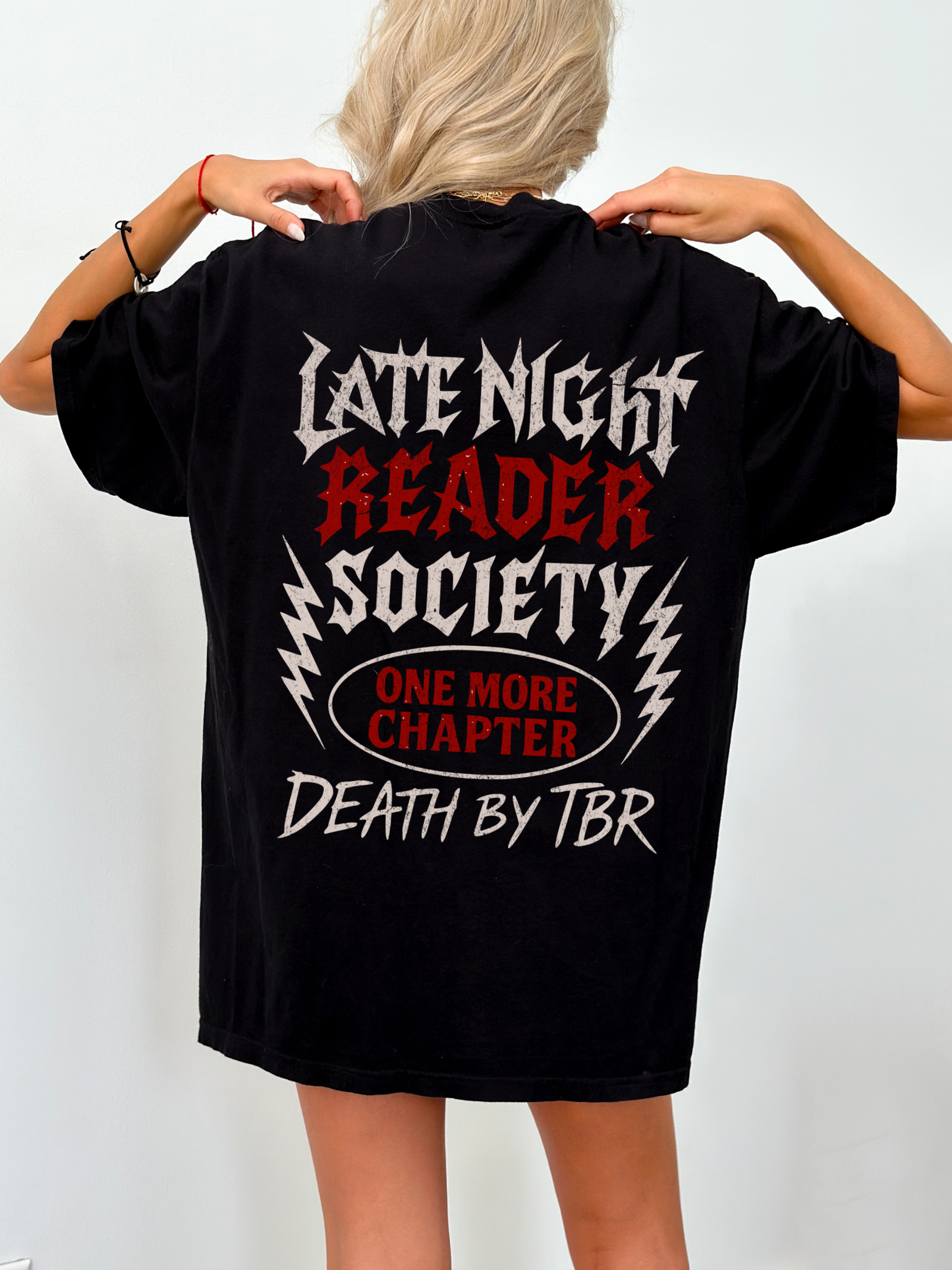 Oversized black t-shirt worn by a blonde woman, showcasing a bold metal-inspired back design with the text “Late Night Reader Society,” “One More Chapter,” and “Death by TBR” in grungy gothic fonts. Ideal gift for fantasy bookworms who stay up reading.