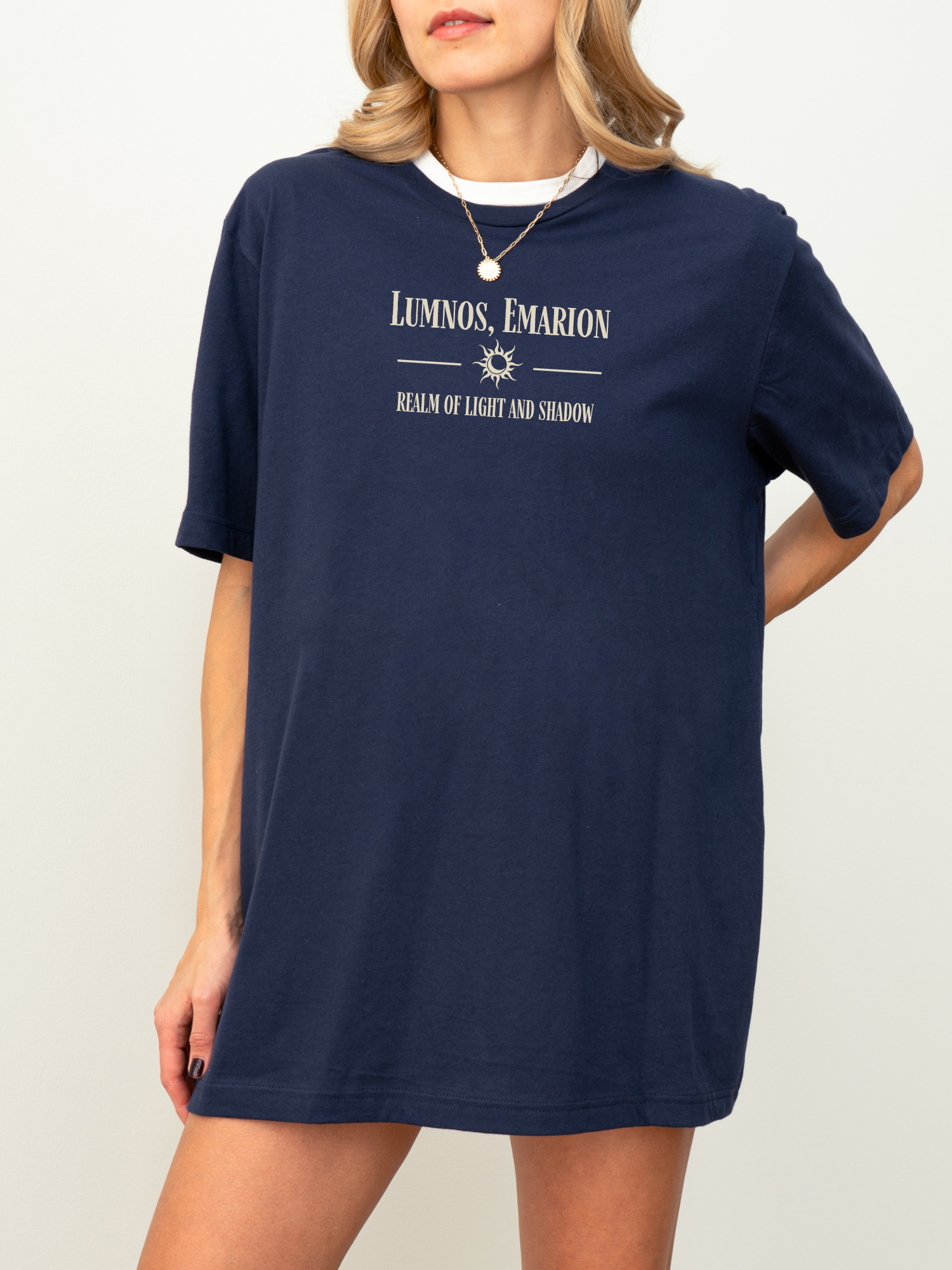 Model wearing a navy blue t-shirt with cream print that reads "Lumnos, Emarion - Realm of Light and Shadow", styled with a gold pendant and white underlayer, standing against a white backdrop