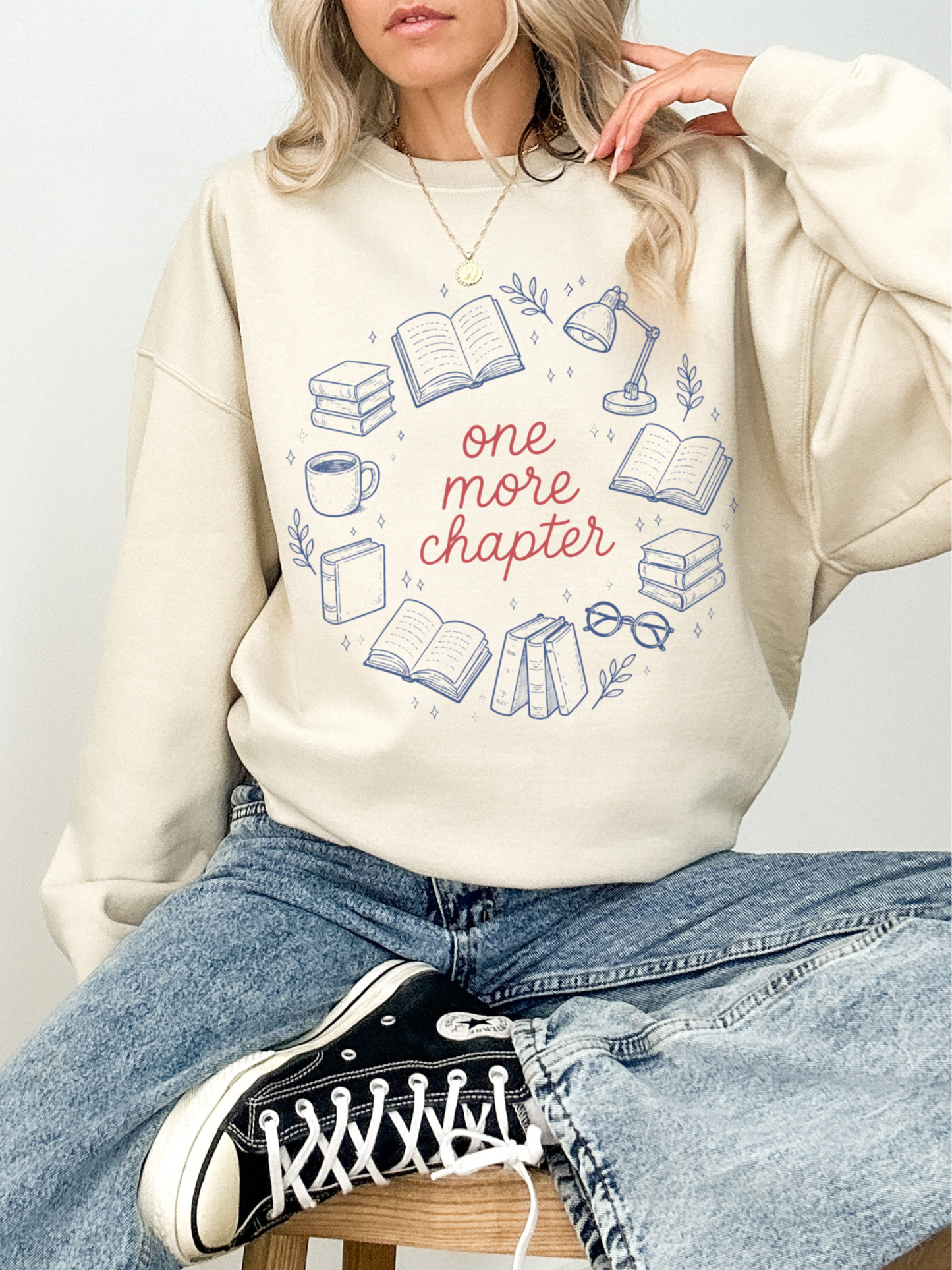Woman sitting casually wearing sand sweatshirt with one more chapter design featuring book illustrations styled with relaxed jeans and sneakers