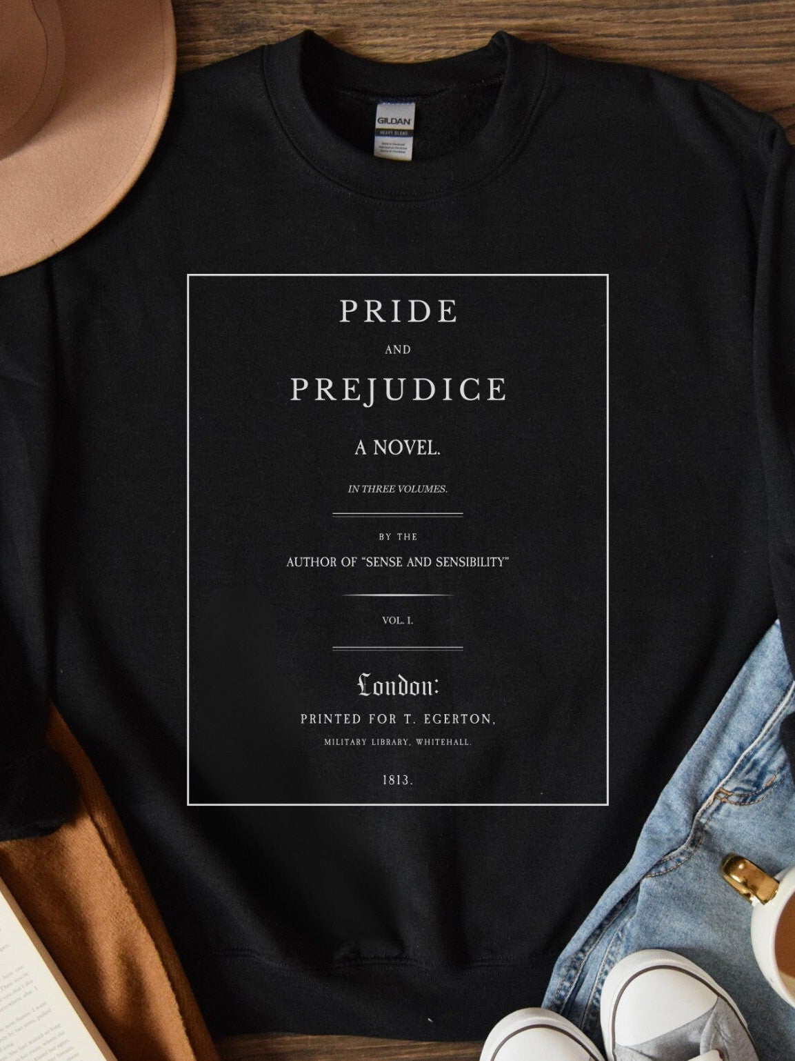 folded black sweatshirt with white Pride and Prejudice title page design styled with jeans and hat