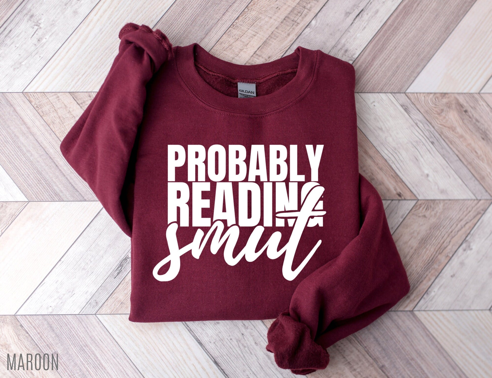 folded maroon Probably Reading Smut sweatshirt with large white bold text on wood floor