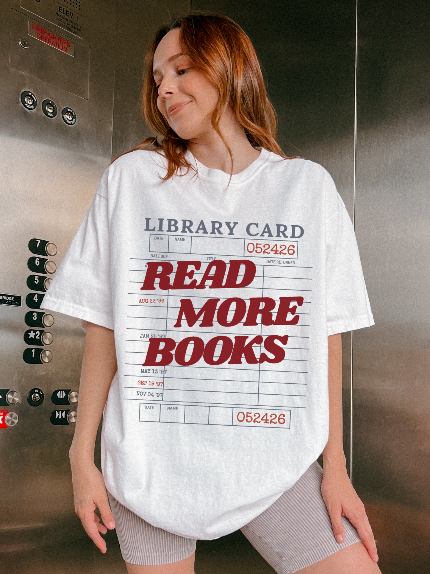 woman wearing a white Comfort Colors shirt featuring a vintage library card read more books design in an elevator setting, oversized reader graphic tee perfect for librarians bookworms and reading enthusiasts