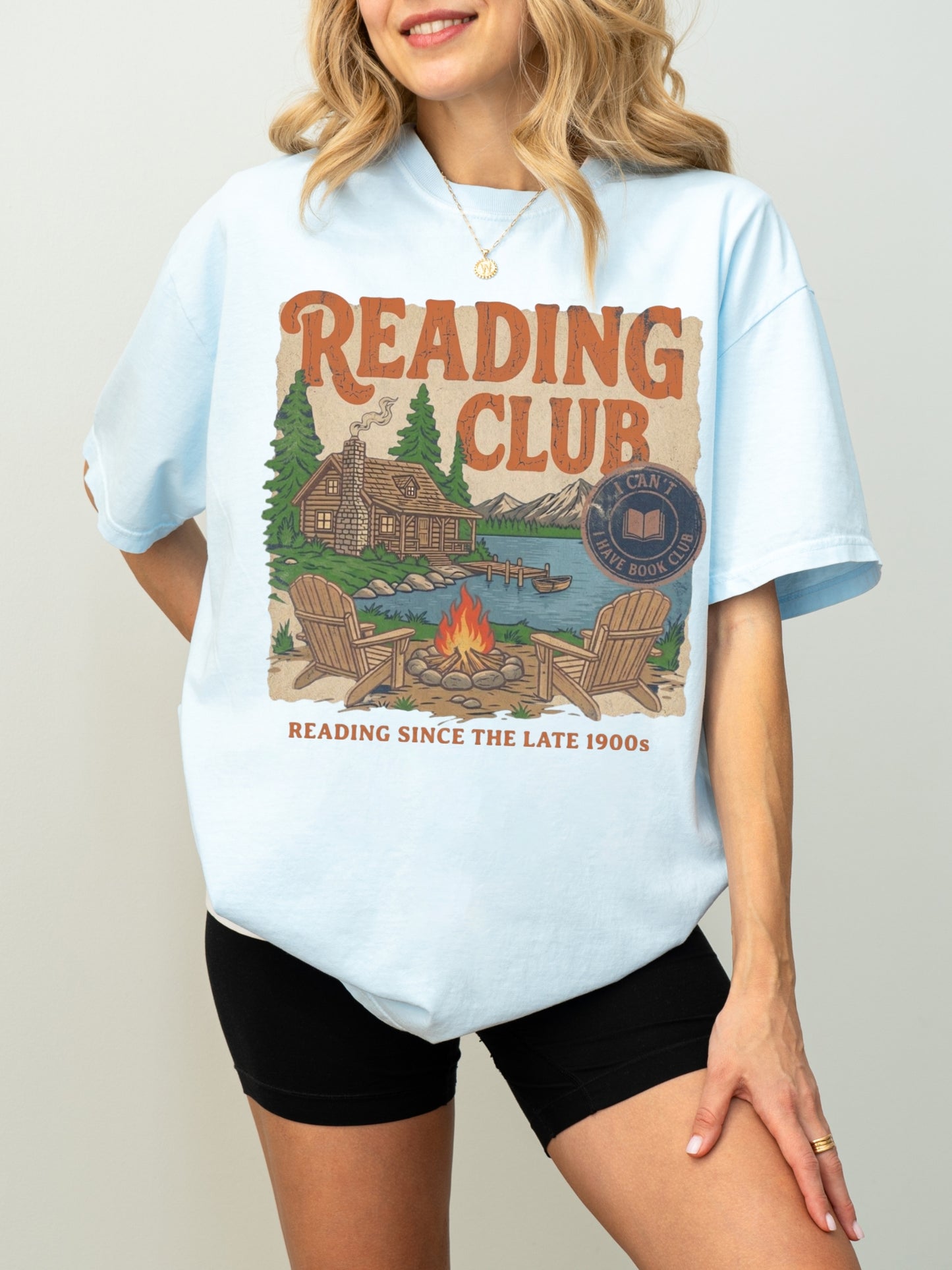 A woman wearing an oversized chambray graphic T-shirt with a vintage-style 'Reading Club' print on the front, paired with black bike shorts, against a plain light gray studio background.