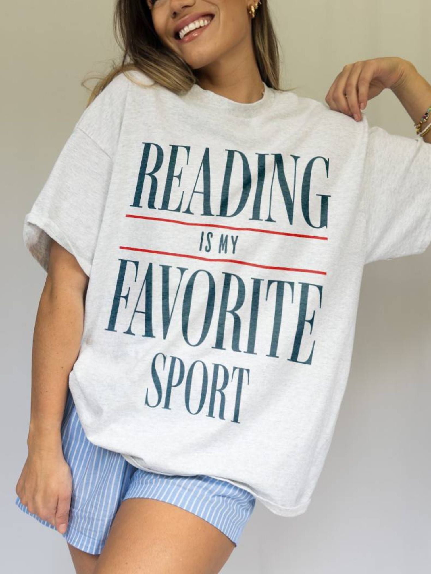 Woman wearing oversized "Reading Is My Favorite Sport" graphic tee in light grey with bold text, smiling casually.