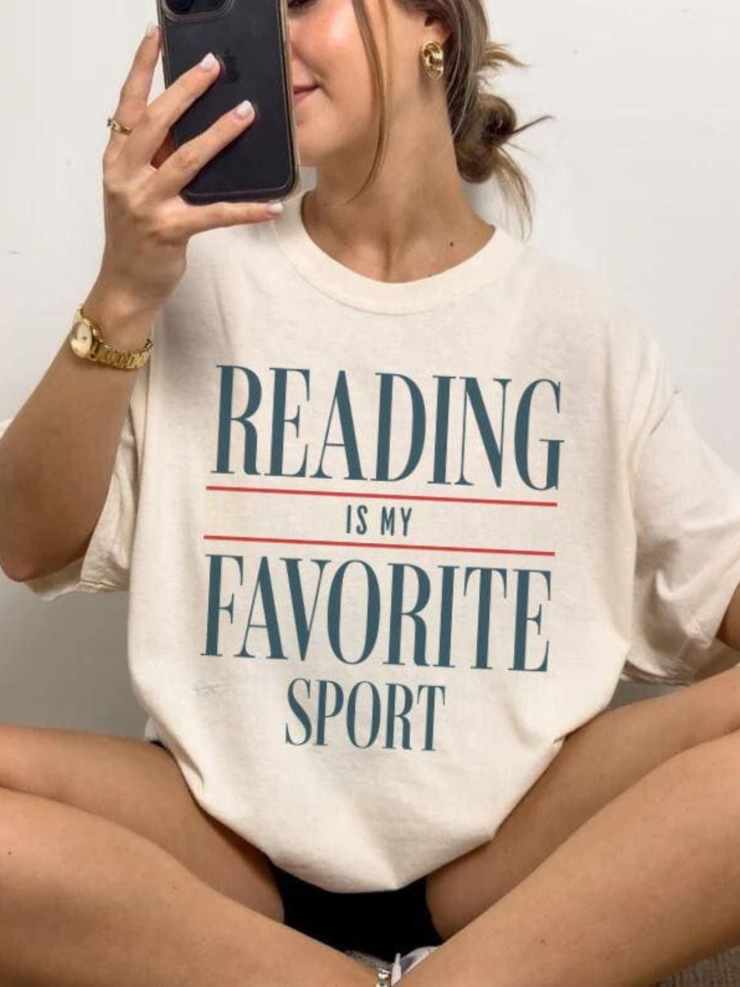 Woman wearing oversized "Reading Is My Favorite Sport" graphic tee in natural color with bold text, smiling casually taking a selfie with her phone