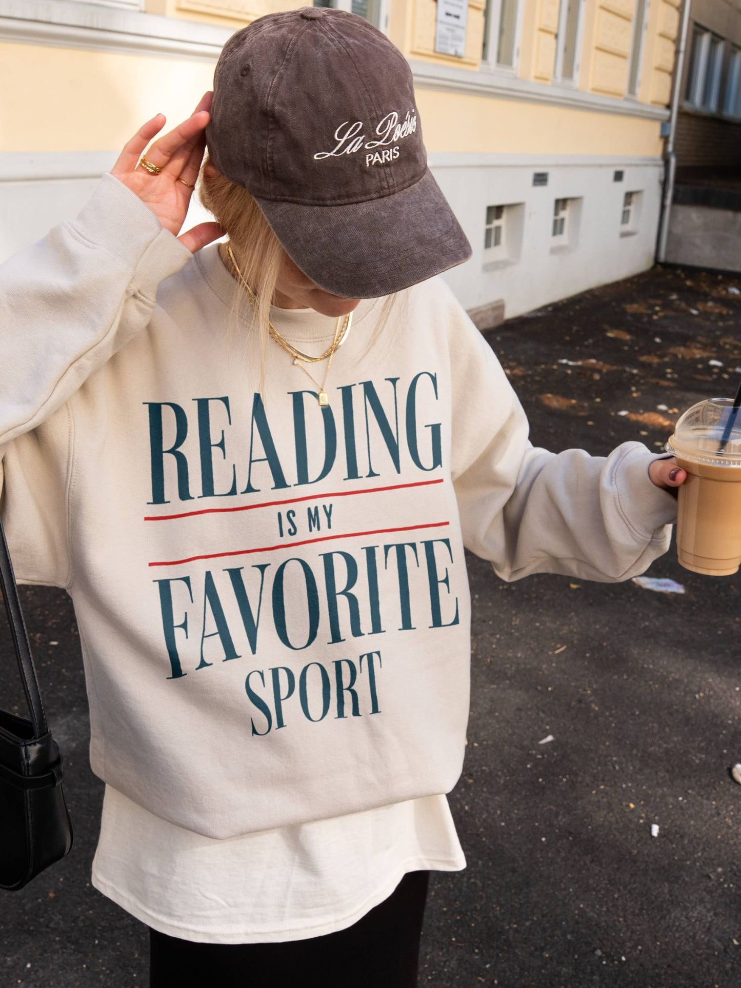 woman outside wearing a sand-colored sweatshirt that says reading is my favorite sport in bold retro text