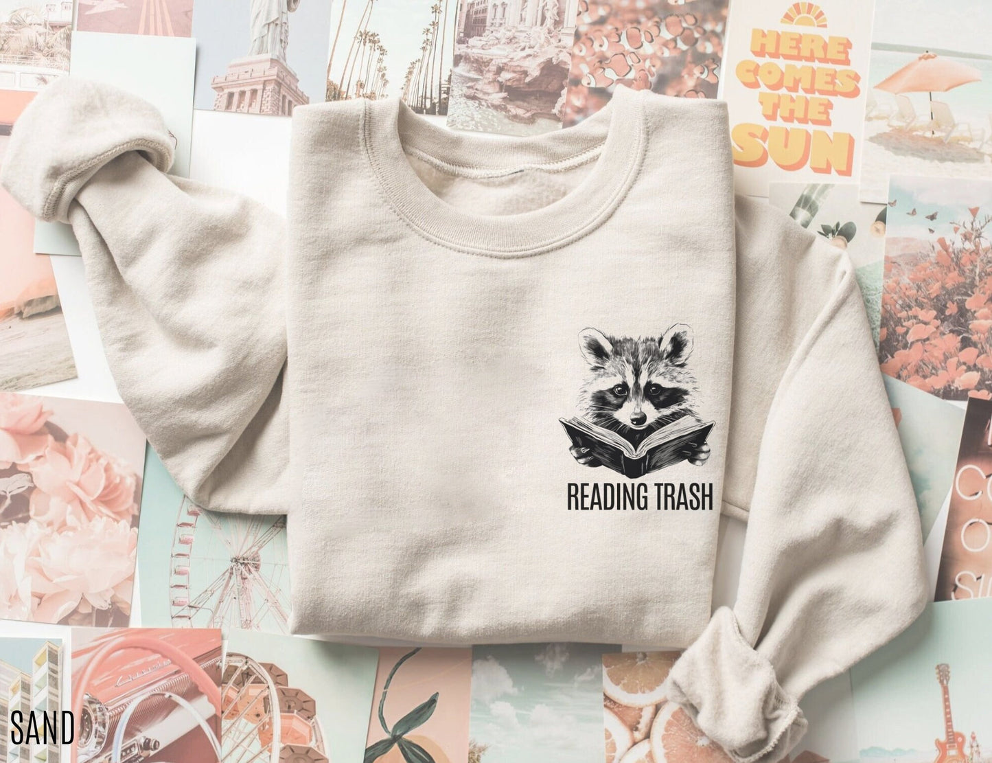 folded sand-colored sweatshirt with black sketch of raccoon reading and text reading trash