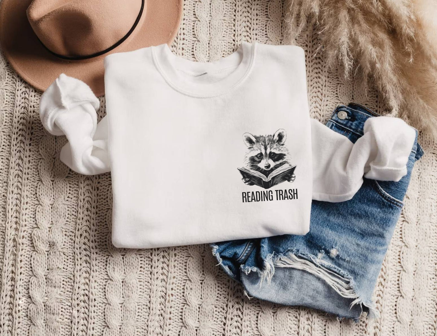 folded white sweatshirt with cute raccoon holding a book and funny reading trash text on front