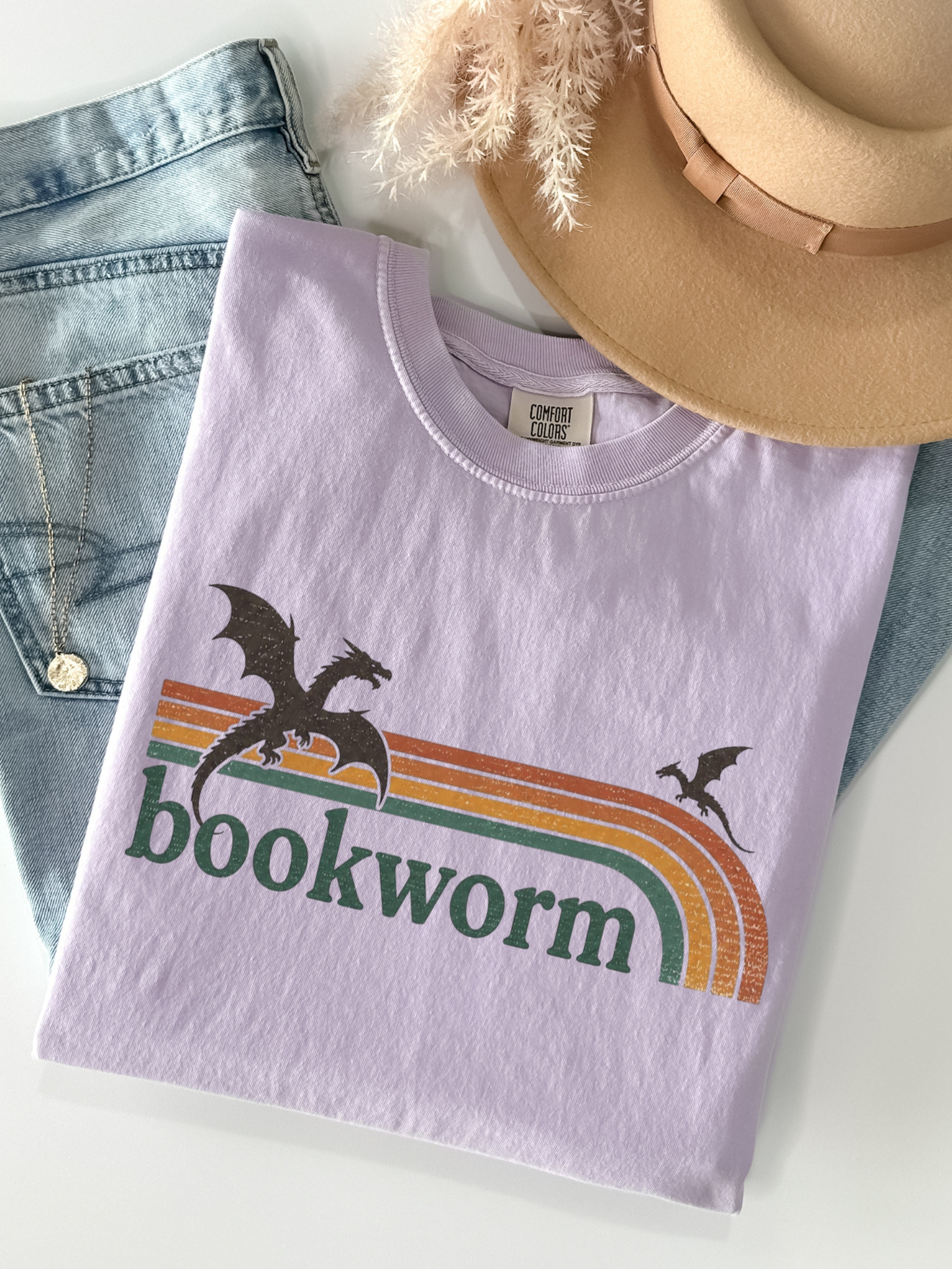 orchid comfort colors bookworm dragon shirt flat lay with retro rainbow graphic styled with jeans jewelry and beige hat