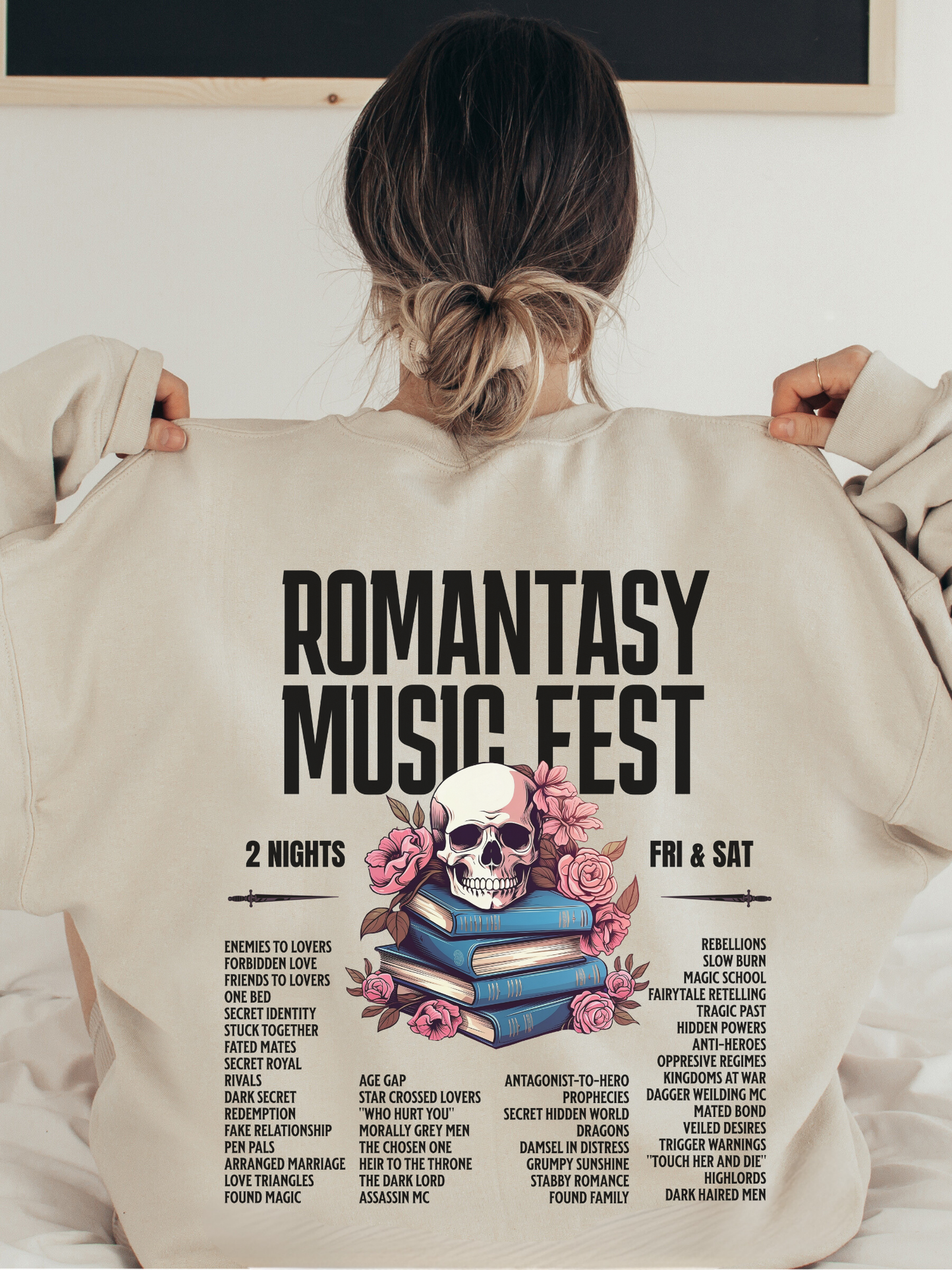 Sand-colored sweatshirt laid flat with the Romantasy Music Fest concert-style graphic on the back, listing iconic romantasy tropes like found family, magic school, and grumpy sunshine alongside a skull and book stack design.