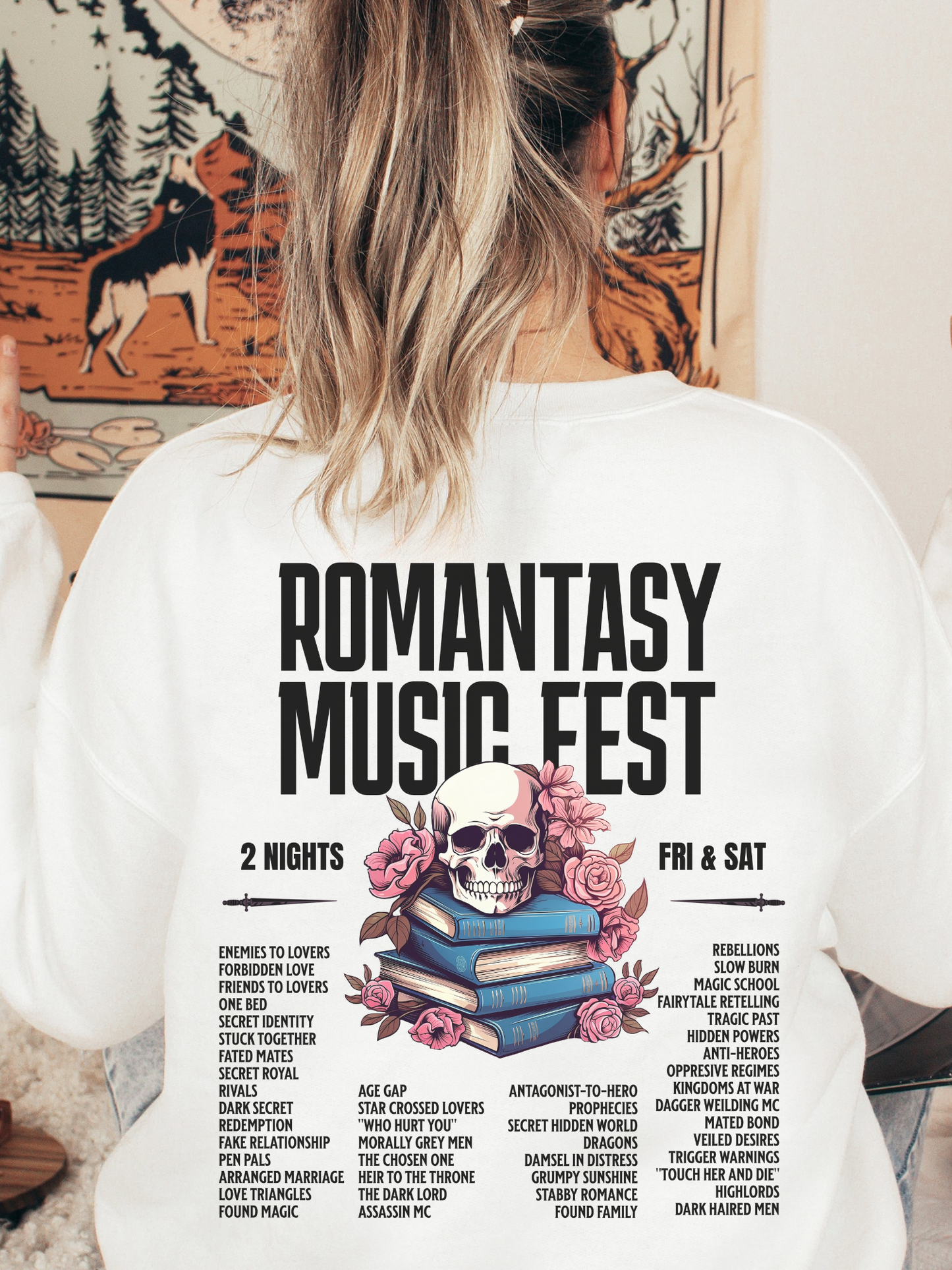 White sweatshirt with Romantasy Music Fest graphic on the back, including a pink floral skull atop books and fantasy tropes like morally grey men and found magic, modeled with loose waves in a cozy indoor setting.