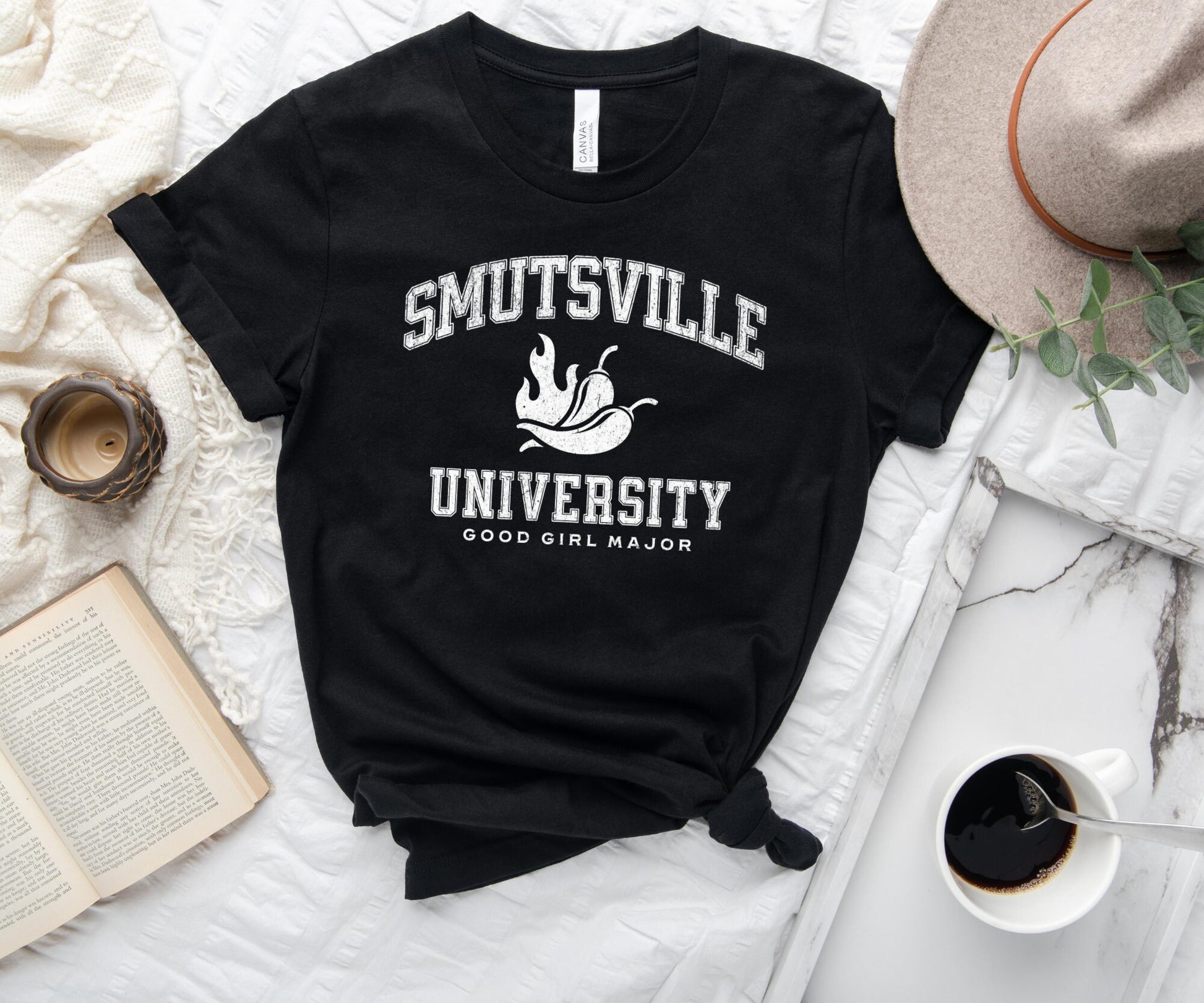 black t-shirt with vintage white Smutsville University print, laid flat with coffee, book, and beige hat