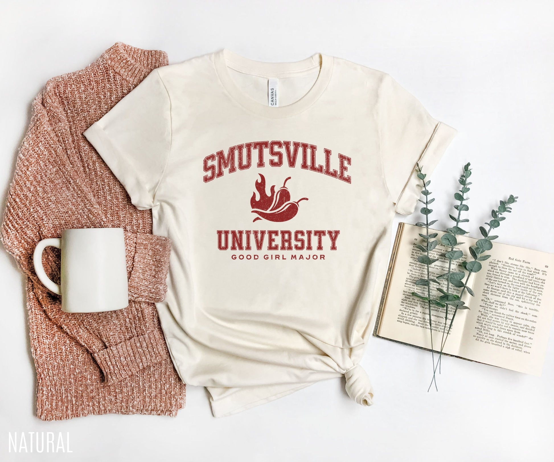 folded natural t-shirt with “Smutsville University Good Girl Major” text in red, styled with rust cardigan, white mug, open book, and eucalyptus