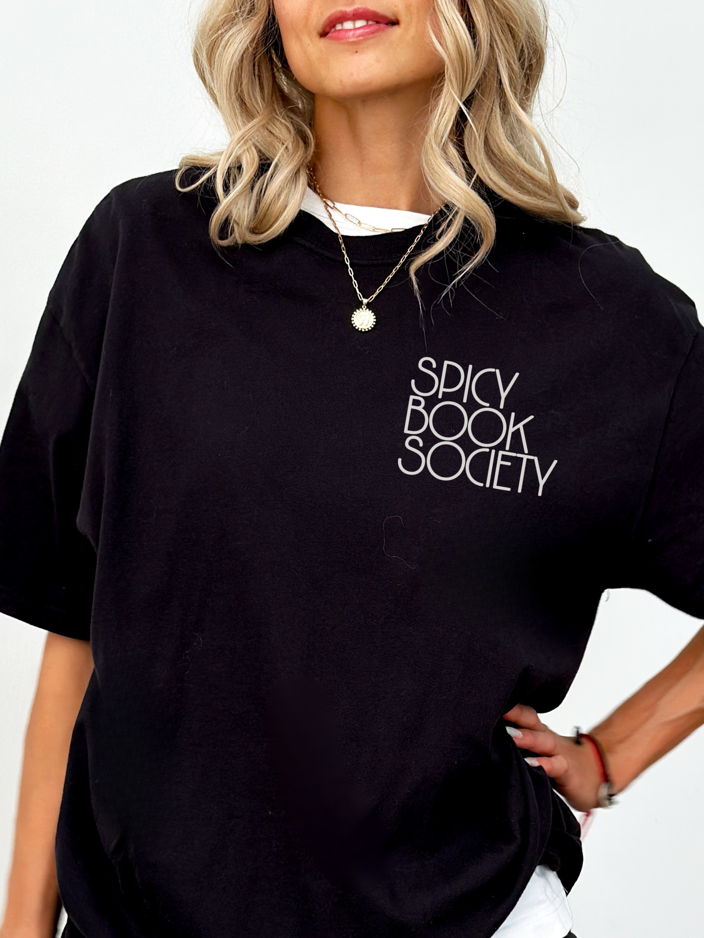 Close-up of a woman in a relaxed-fit black Comfort Colors tee with “Spicy Book Society” on the left chest. Aesthetic reader outfit with cozy book club energy, styled with soft curls and dainty jewelry.