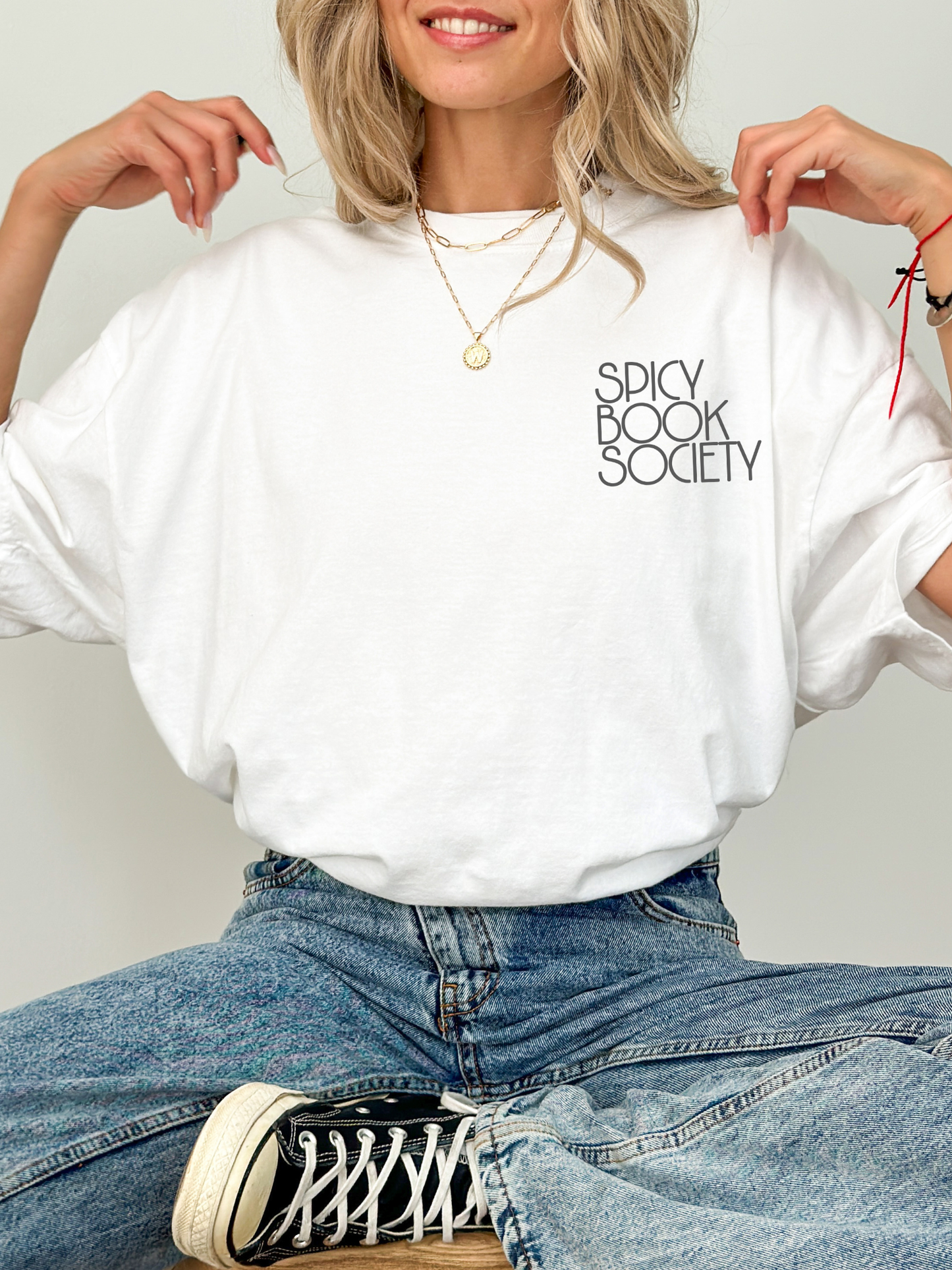 Woman sitting cross-legged wearing a white Comfort Colors T-shirt that says “Spicy Book Society.” Styled with jeans and layered gold necklaces in a cozy, bookish setting perfect for romantasy and smut readers.