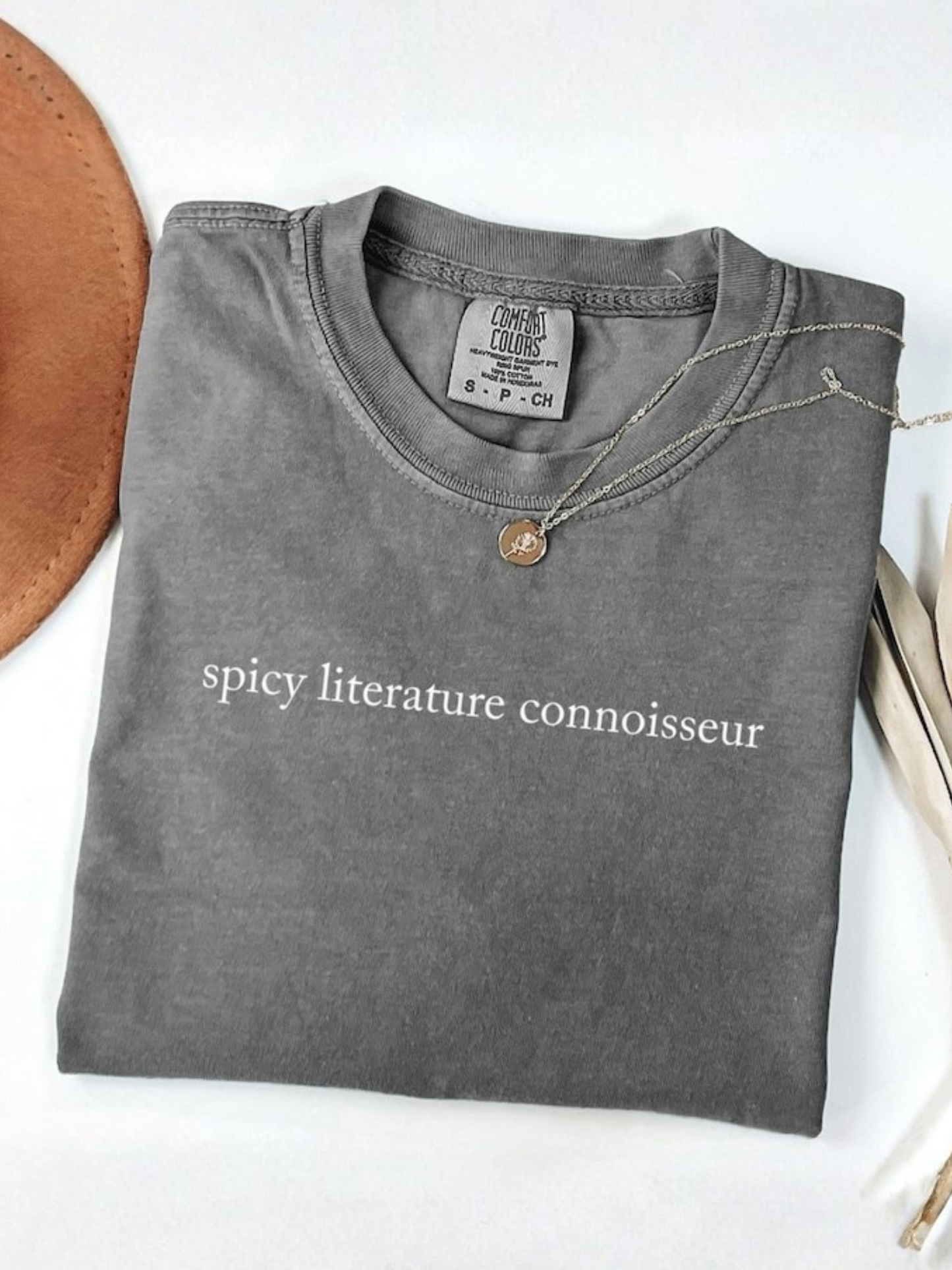 folded grey Comfort Colors shirt with spicy literature connoisseur saying next to neutral hat and dried florals