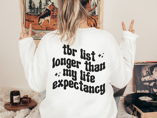 woman wearing white sweatshirt with long TBR list quote flashing peace signs in cozy reading nook