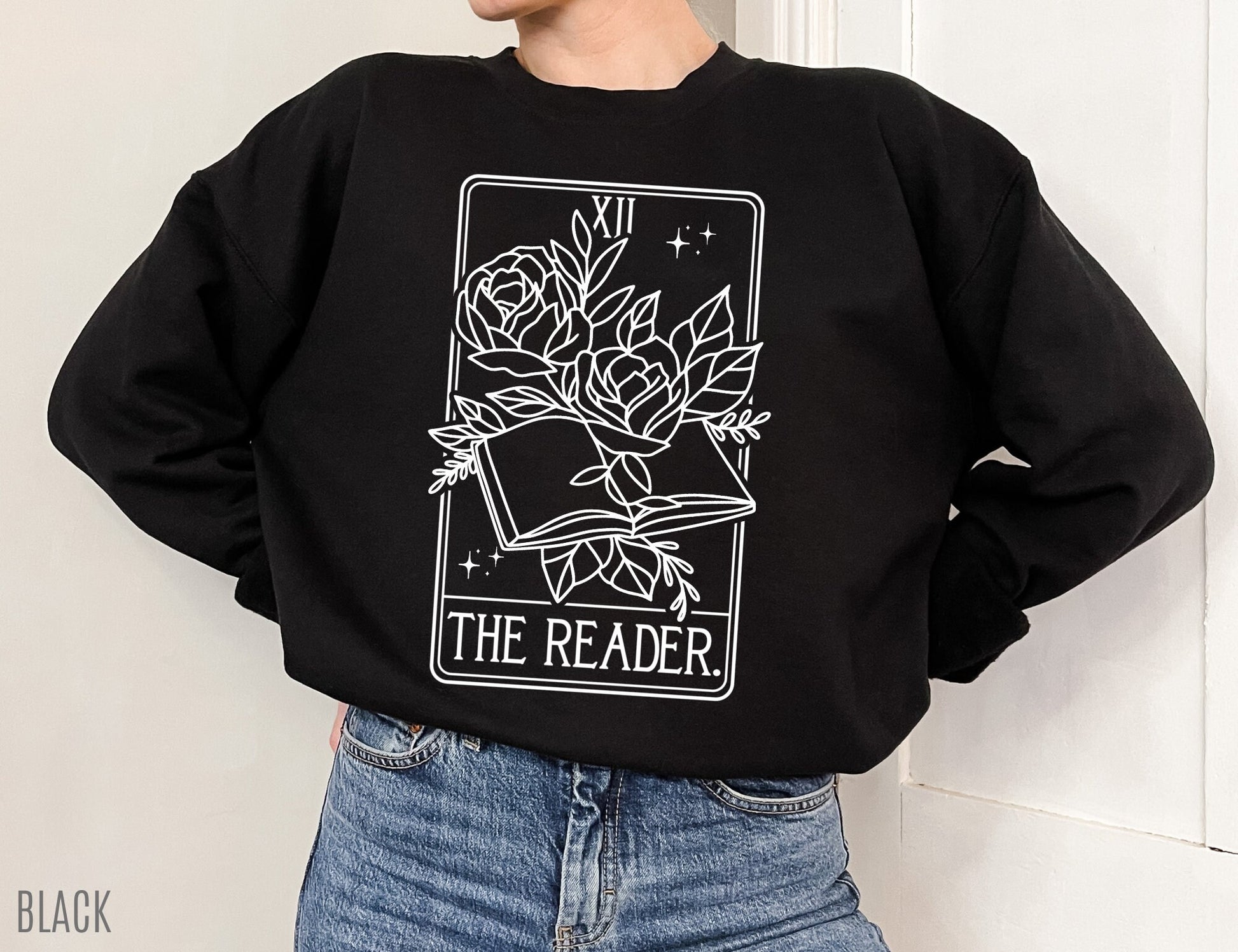 woman in black The Reader tarot sweatshirt with white floral book design and hands in pockets